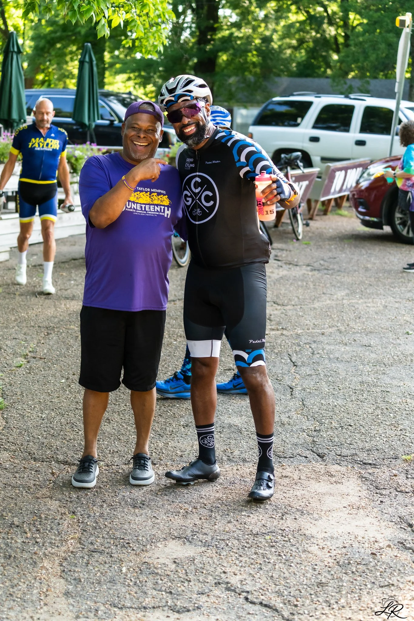Two smiling men standing close together outdoors, with other people and cars in the background. One man is wearing a purple shirt with a bicycle event graphic, shorts, and sneakers. The other man is dressed in cycling gear, including a helmet, sungla