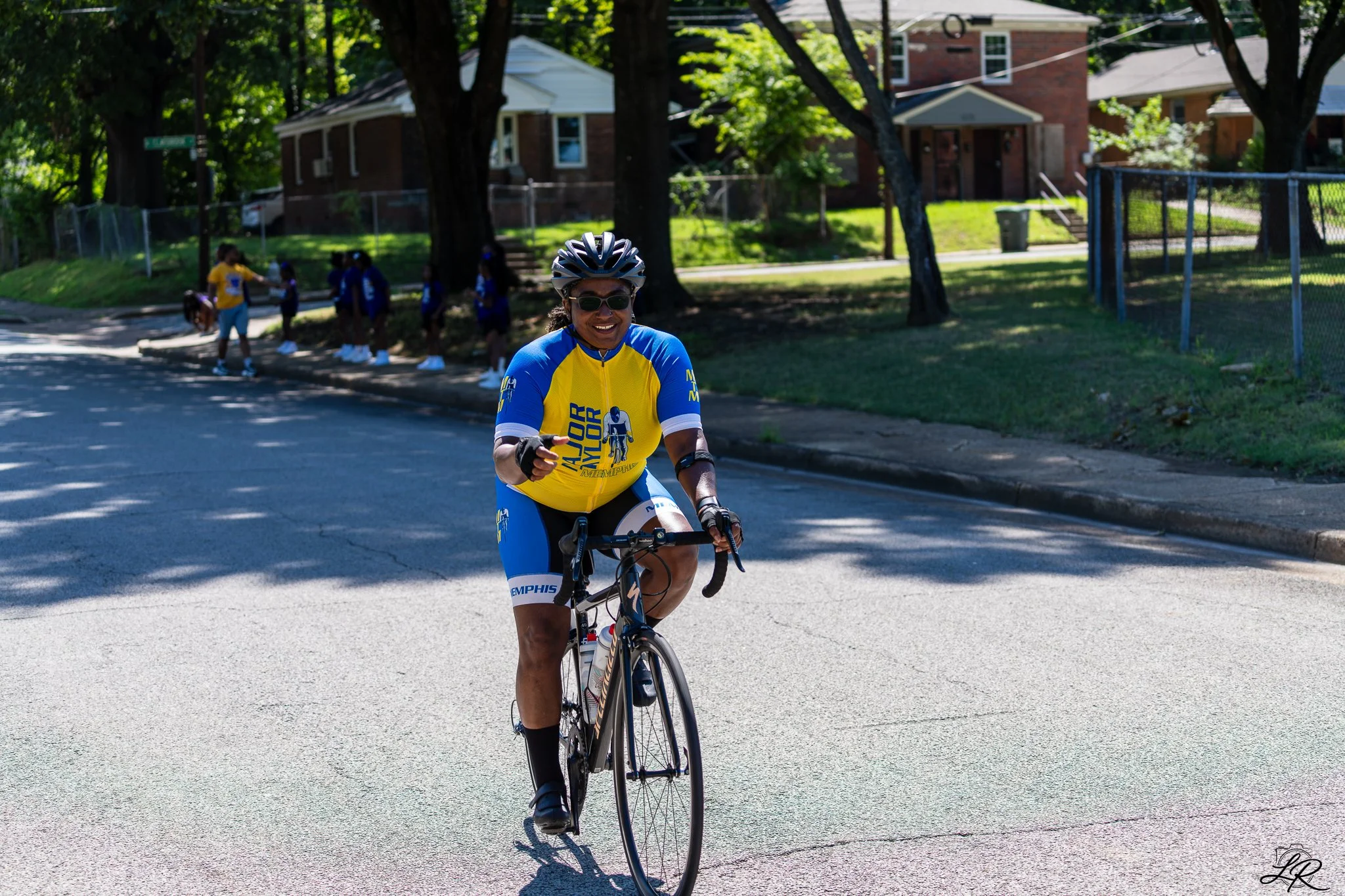 Woman in cycling gear, wearing a yellow and blue shirt, helmet, and sunglasses, riding a bicycle on a sunny street with trees and residential houses in the background.