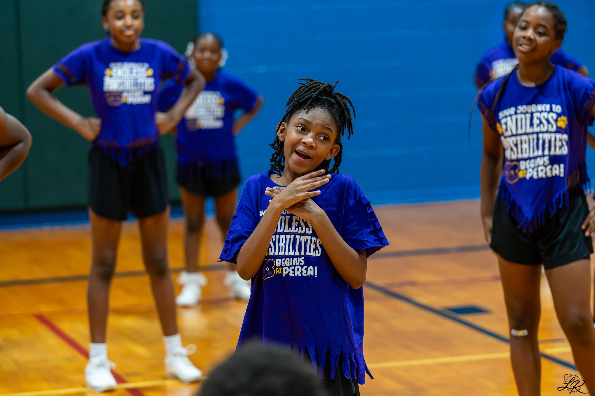 A group of young children participating in a sports activity in a gymnasium, wearing purple shirts with the phrase 'Endless Possibilities' printed on them. The girl in the foreground is gesturing with her hands and smiling, surrounded by other childr