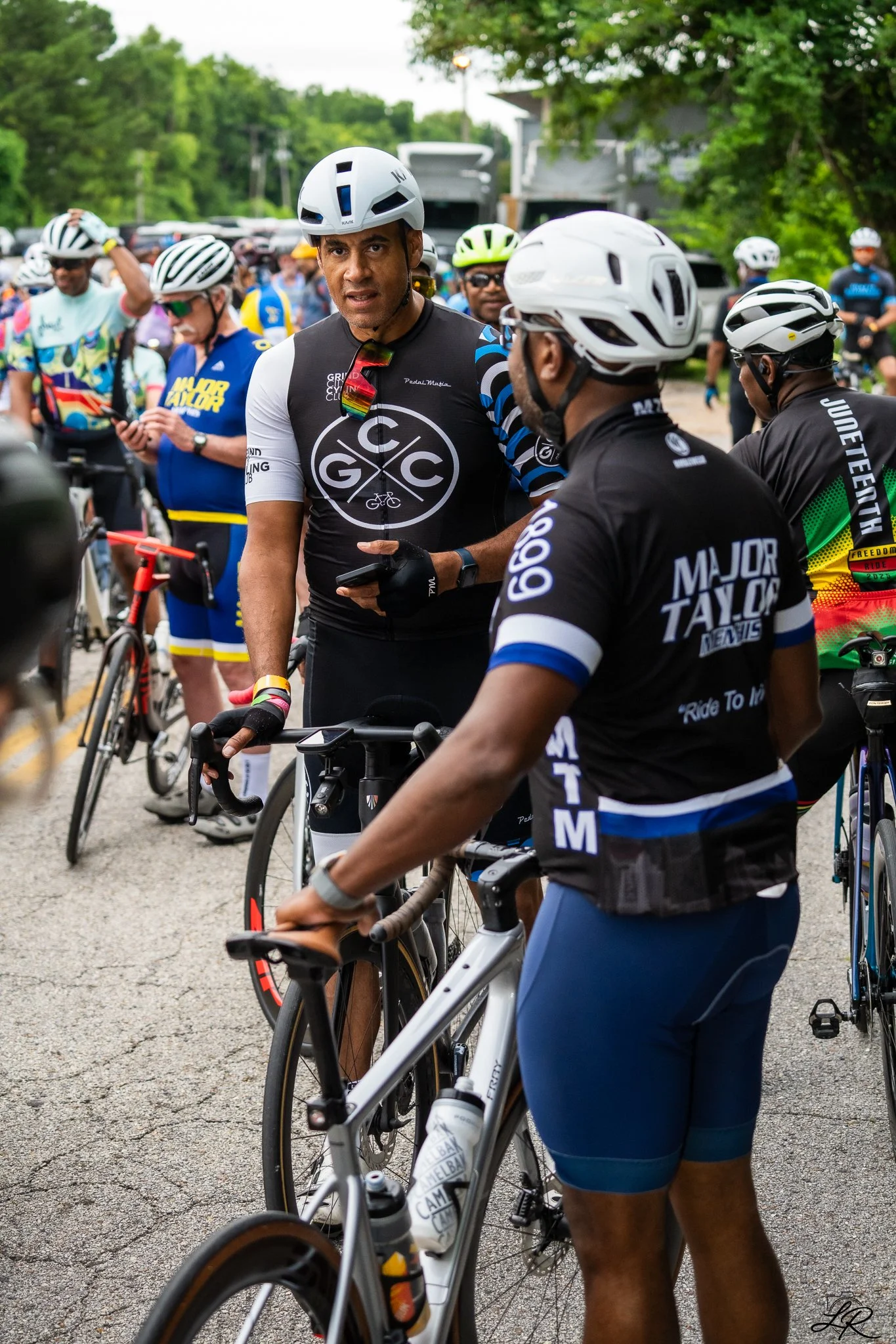 A group of cyclists wearing helmets and cycling gear gathered outdoors on a paved area, with trees and parked cars in the background, preparing for or finishing a bike ride.
