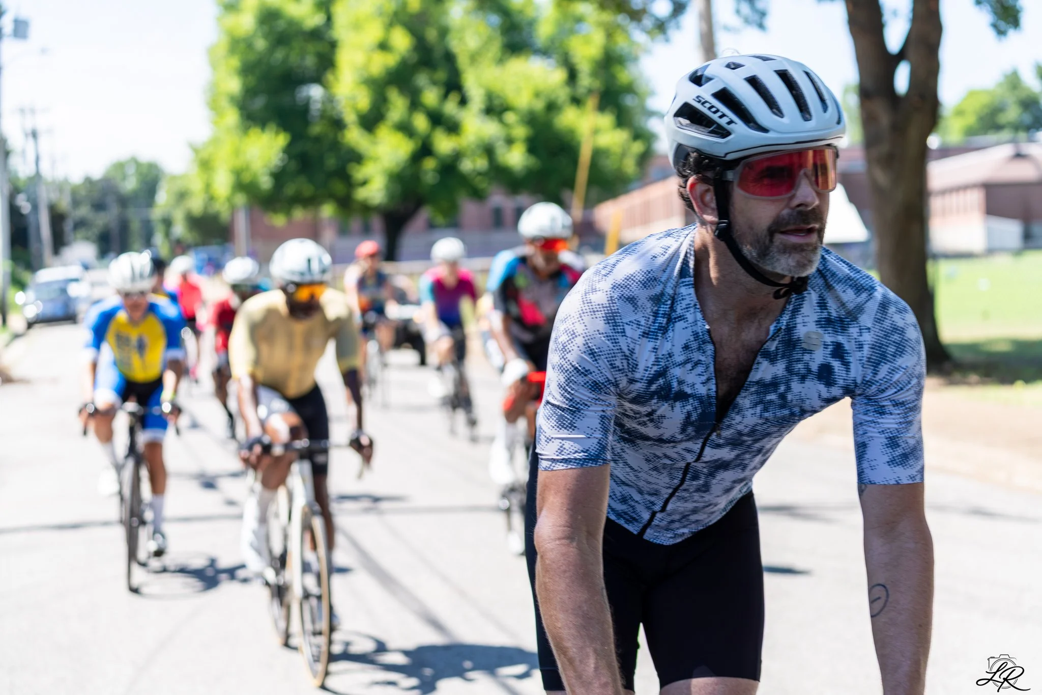 A group of cyclists riding on a sunny street with trees and houses in the background. The foreground features a man with a helmet and sunglasses, wearing a patterned cycling jersey.