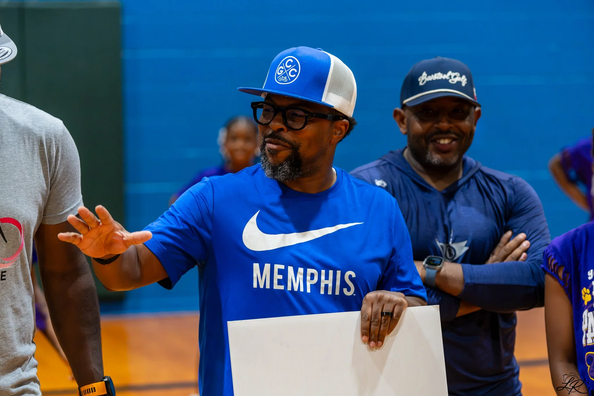 Two men smiling in athletic clothing, one wearing a blue Memphis sports shirt and a blue and white cap, and the other a dark blue cap, standing in an indoor sports facility.