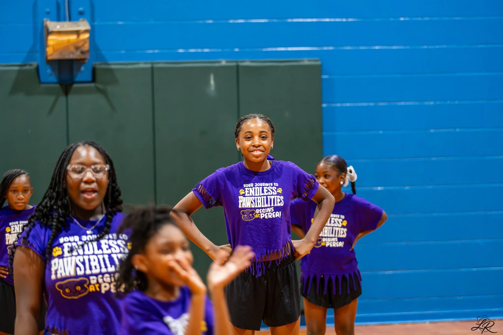 Young girl in purple shirt smiling confidently with her hands on her hips, surrounded by other children in a gymnasium.