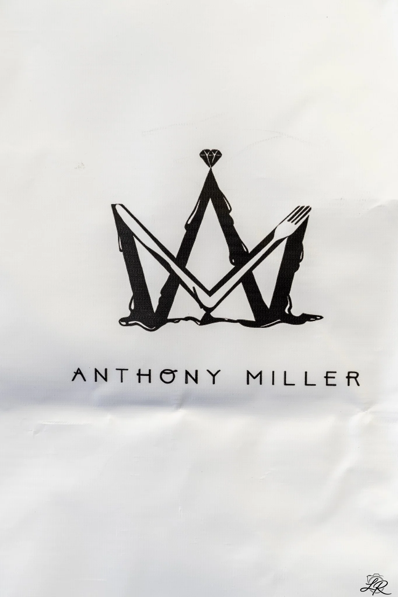 Book cover with a minimalist black and white design featuring a fork, knife, and spoon arranged to form a letter 'M' with a diamond on top. The title is 'Antony Miller' written below in black letters.