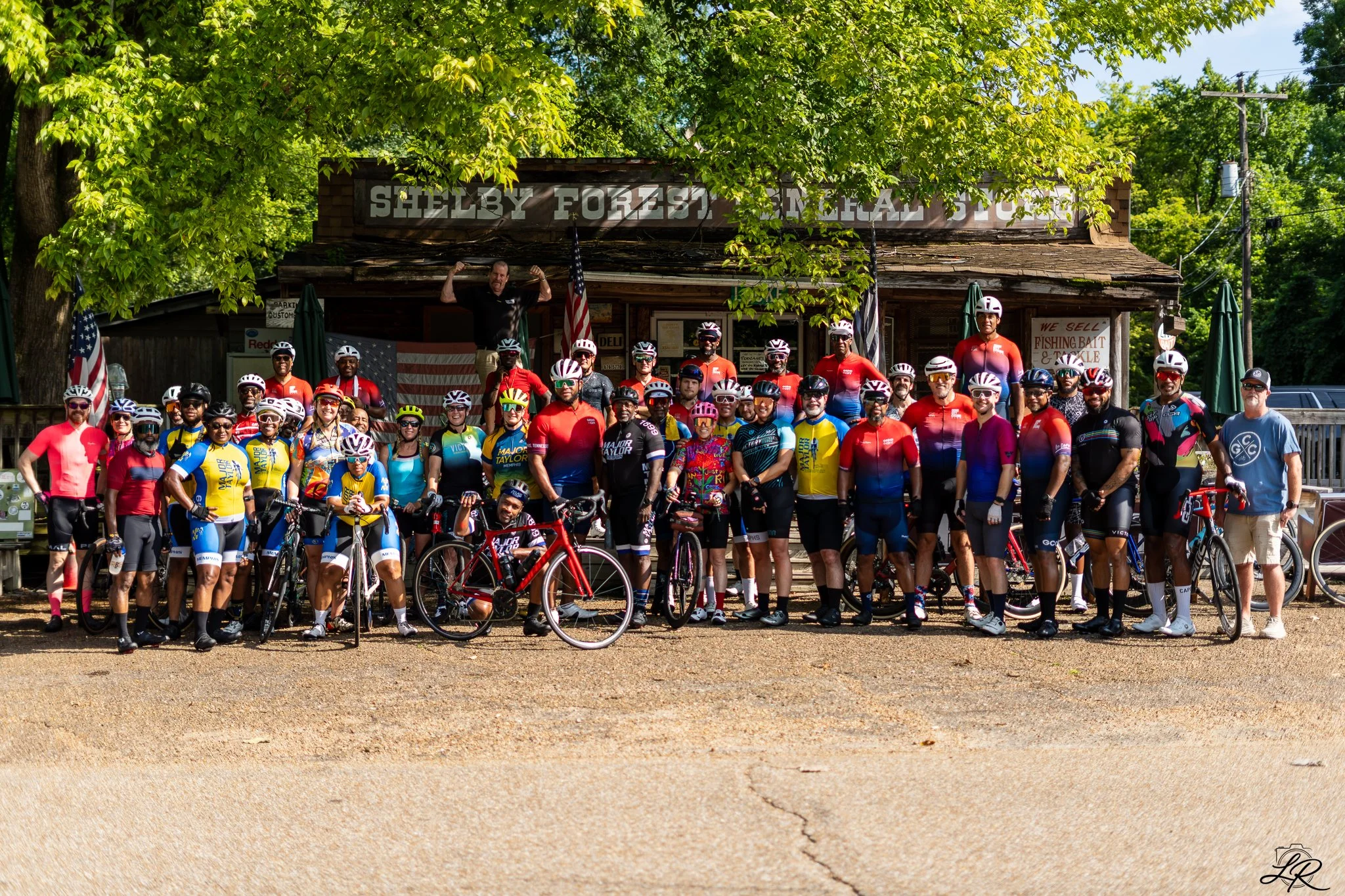 Group of cyclists with helmets and colorful clothing posing in front of a rustic building with a sign that reads 'Shelby Forest General Store.' Some individuals are holding bikes, and there is a man standing on the porch flexing his arms with an Amer