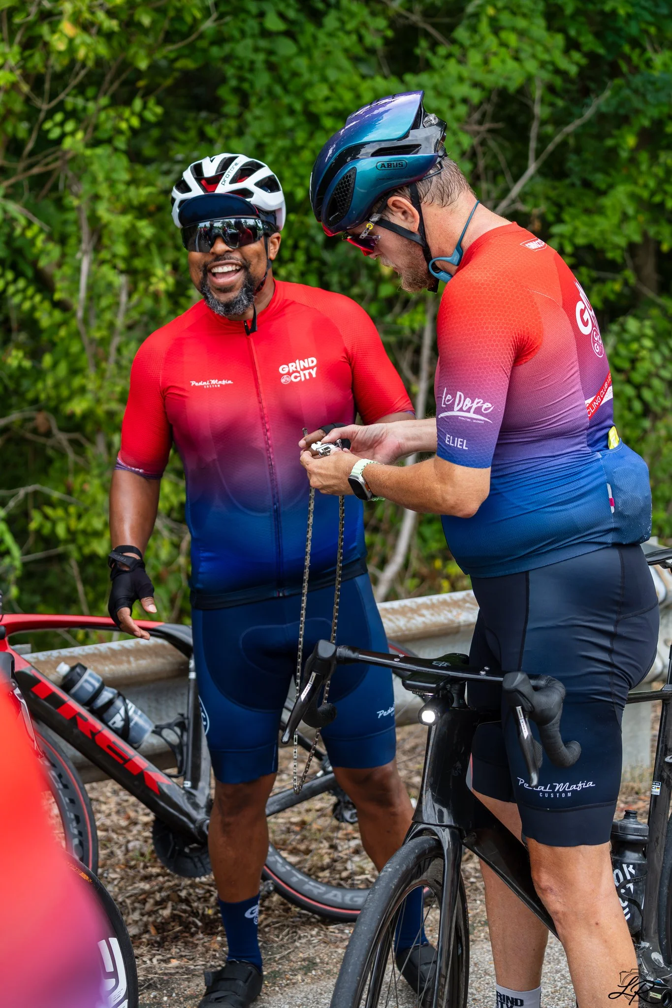 Two men in cycling gear, one smiling and the other looking down at his phone, standing with their bikes outdoors in front of green foliage.