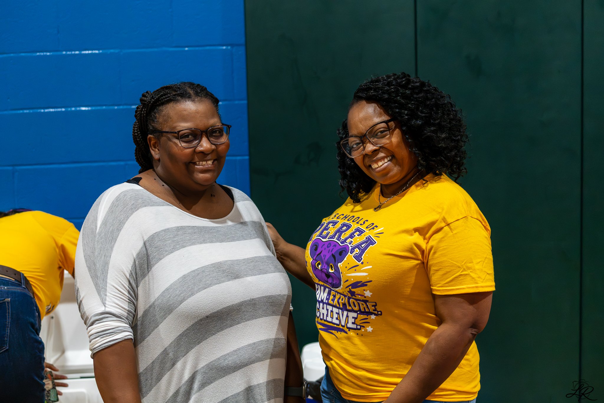 Two women smiling and standing close together indoors, with a blue and green wall in the background. The woman on the left is wearing a gray and white striped shirt and glasses. The woman on the right is wearing a yellow T-shirt with purple text and 