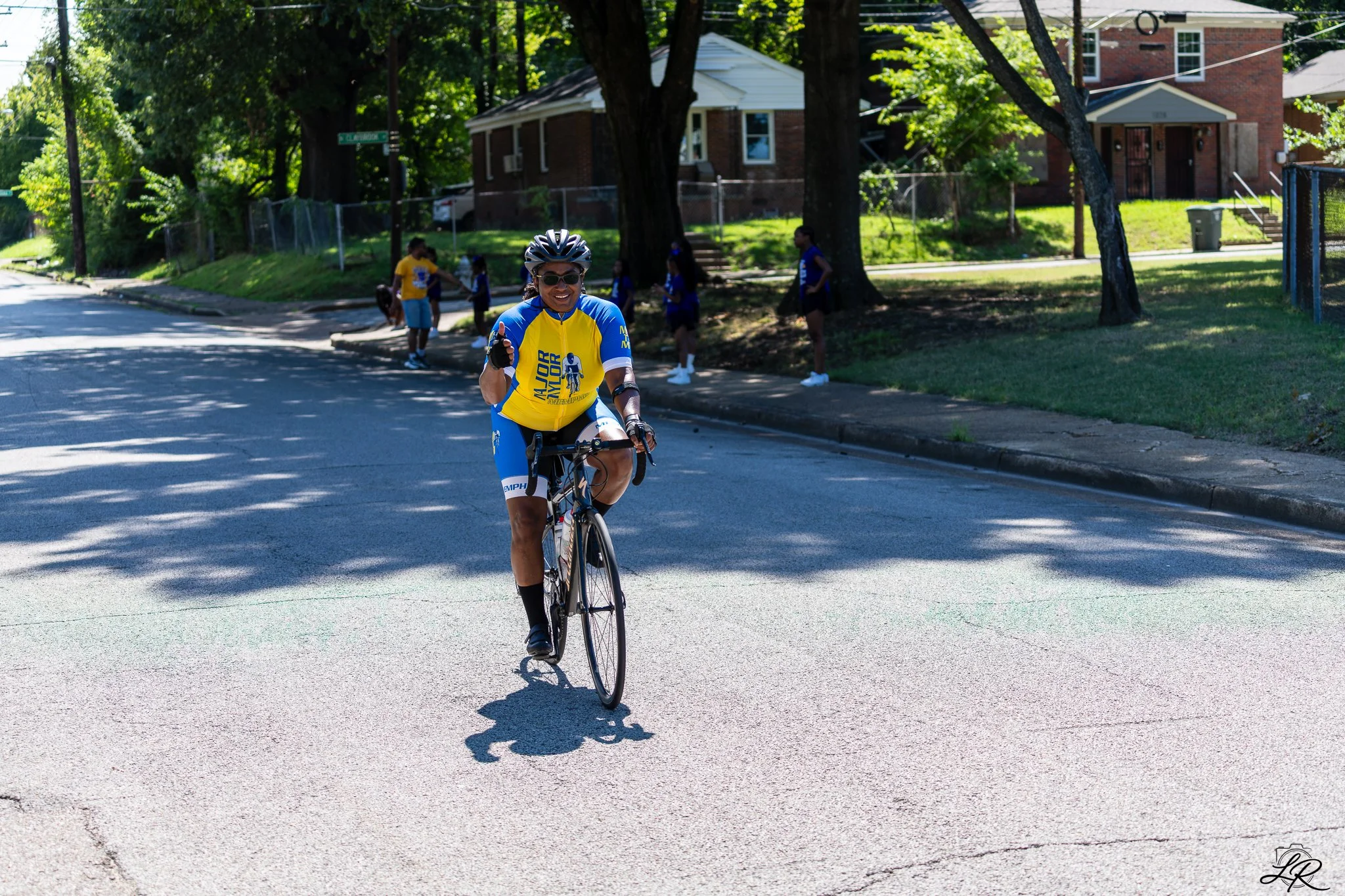 A woman in cycling gear and sunglasses riding a bicycle on a suburban street, with several people in the background, trees, and houses.