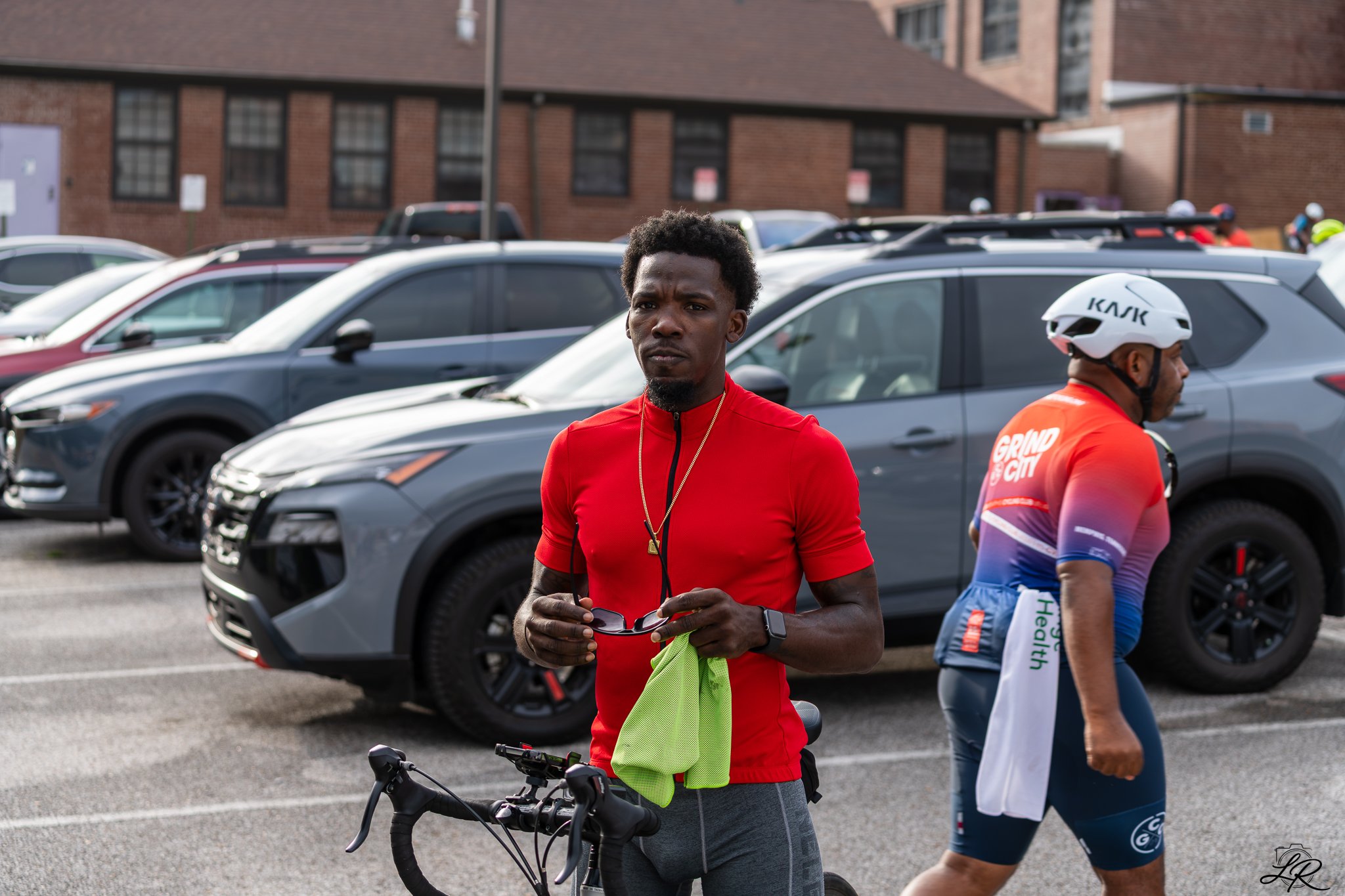 A man with dark curly hair wearing a red shirt holding a small item and standing with a bicycle in a parking lot. A cyclist in colorful attire with a helmet is nearby, and there are multiple parked cars and a brick building in the background.