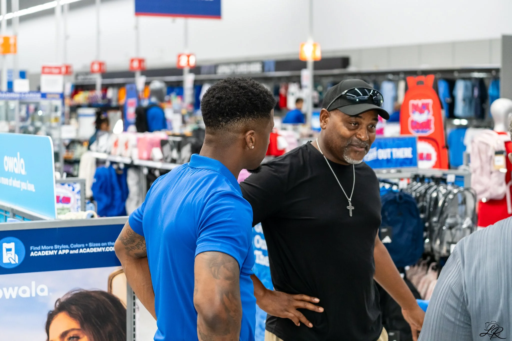 A young man in a blue polo shirt with tattoos on his arm, and an older man wearing a black shirt, black hat, and sunglasses, are talking in a retail store. The background shows clothing racks and store signage.