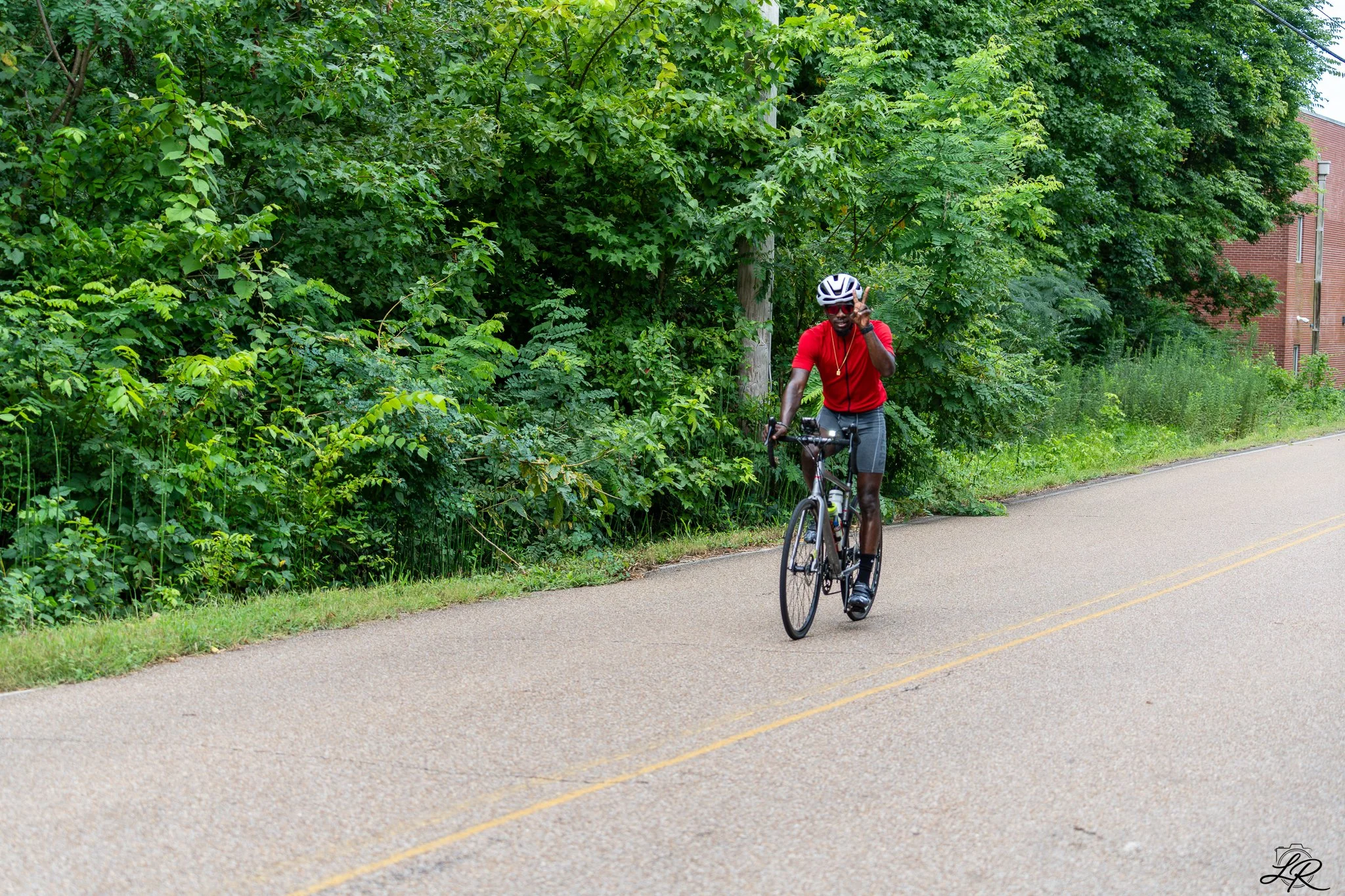 A man wearing sunglasses, a red shirt, gray shorts, and a white safety helmet riding a bicycle along a paved road with yellow lane markings, lined with green trees and bushes, and a brick building visible in the background.