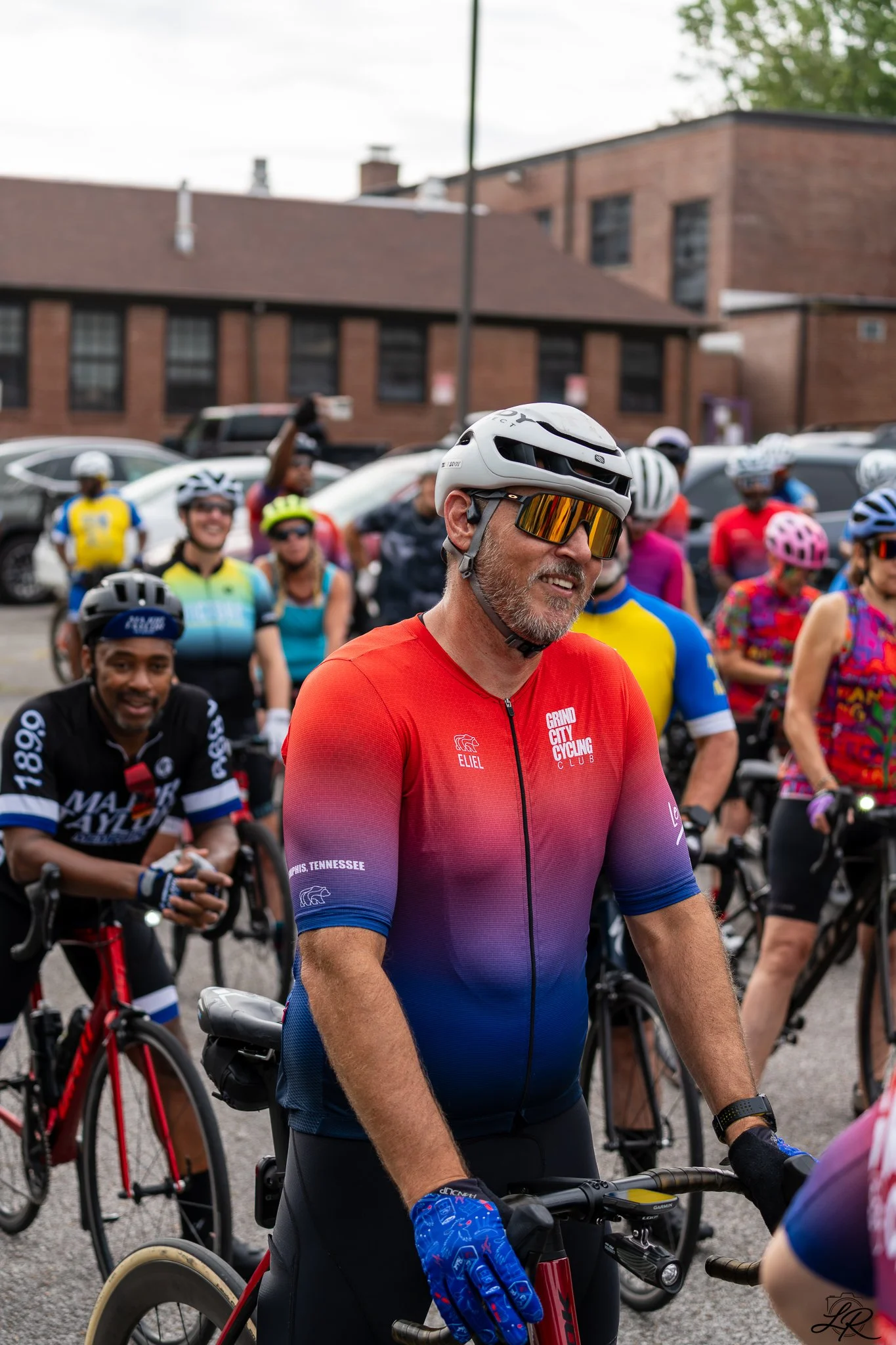 A group of cyclists gathered outdoors, with a man wearing a white bike helmet and reflective sunglasses in the foreground, smiling. The background includes a brick building and several parked cars.