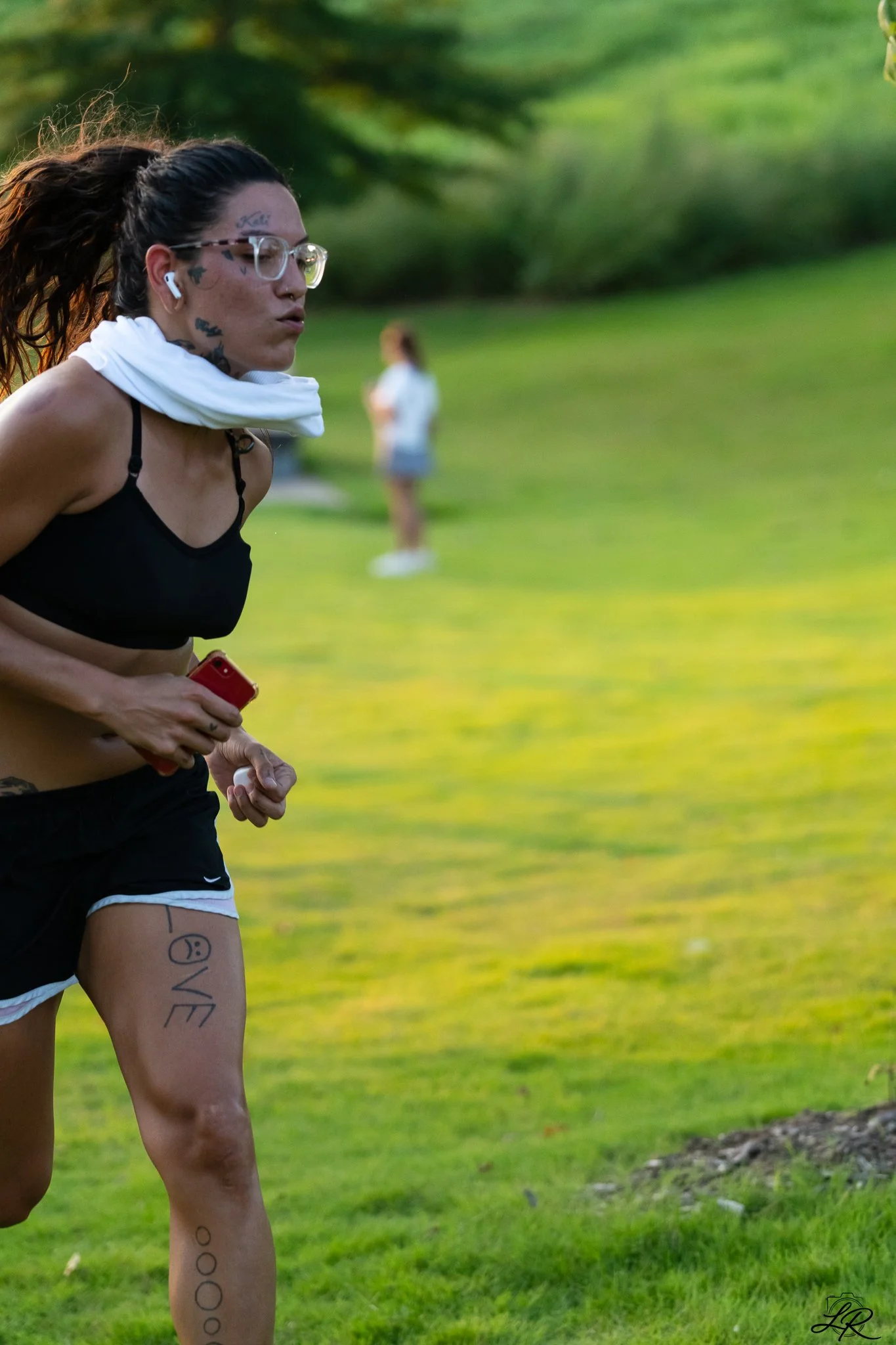 A woman jogging on a grassy field, holding a red phone, with tattoos on her leg, wearing glasses and wireless earbuds, and a white towel around her neck, with trees and another person in the blurred background.