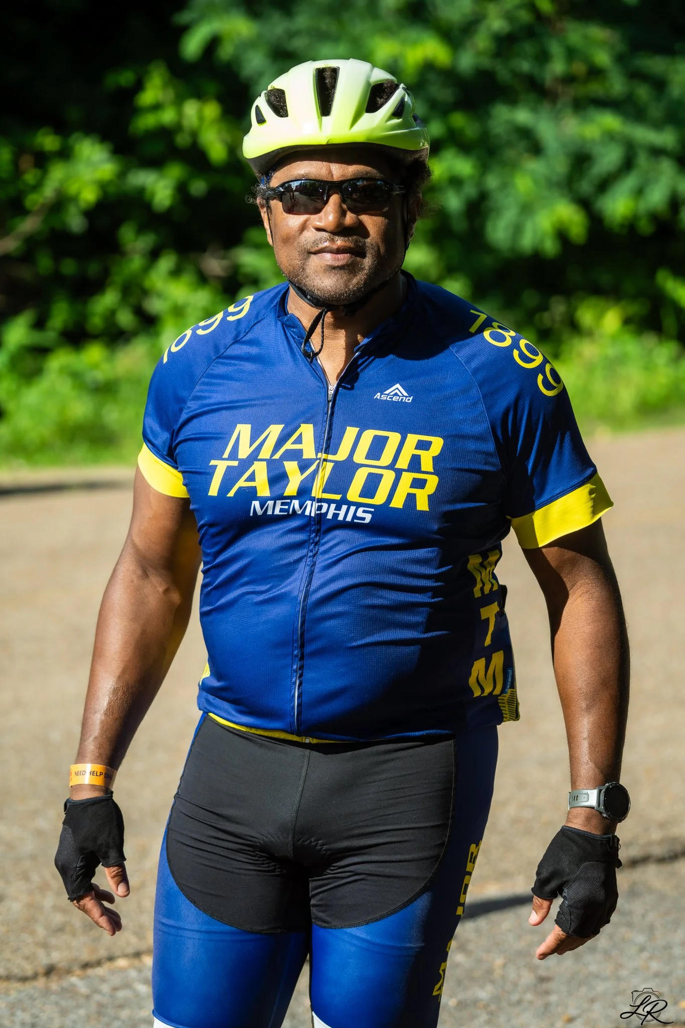 A man standing outdoors on a sunny day, wearing a yellow helmet, dark sunglasses, a blue and yellow cycling jersey with 'Major Taylor Memphis' written on it, black gloves, and sports watch, with greenery in the background.