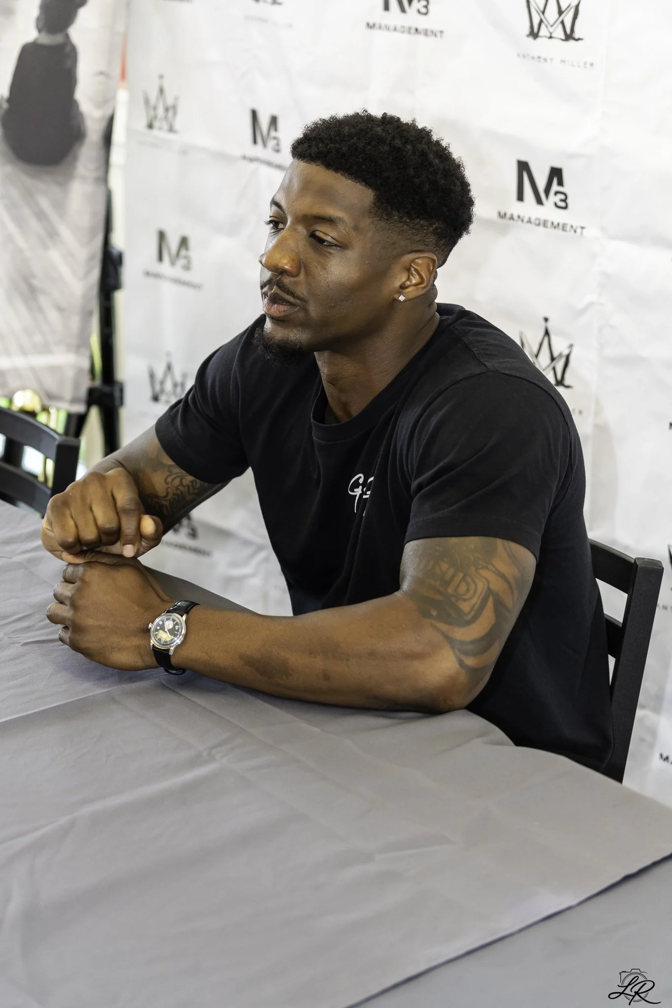 A young African American man with short curly hair and a beard, wearing a black T-shirt, is sitting at a table with his arms resting on it, clasped together. He has a wristwatch on his left wrist and diamond earrings. The background features a white 