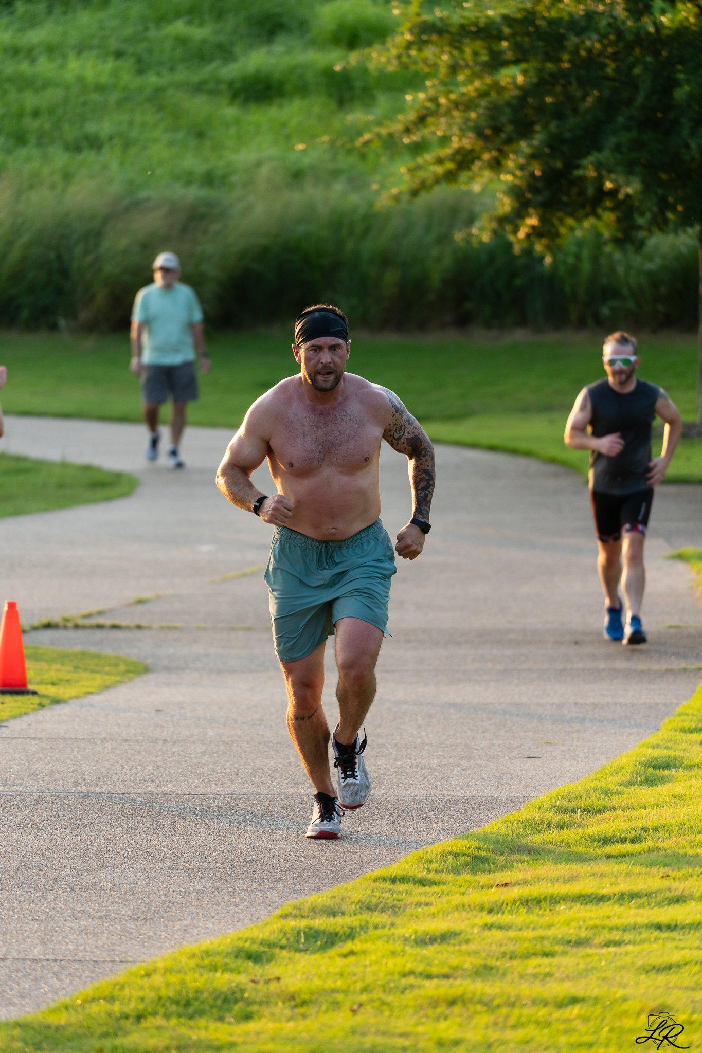 A group of runners participating in a race outdoors on a paved path, with lush green grass and trees in the background, during what appears to be late afternoon or early evening.