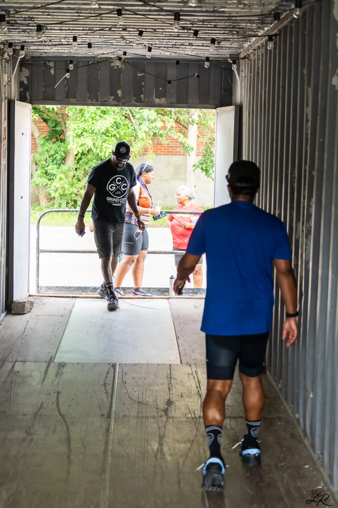 People walking out of a garage or shed, with some wearing rollerblades, amidst green trees and a brick wall in the background.