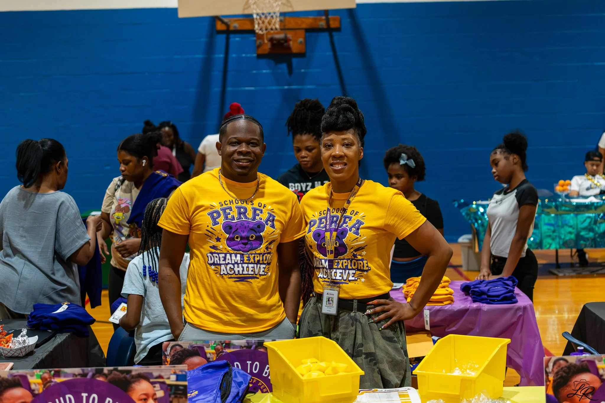 Two women wearing yellow shirts with a lion logo and the words "Schools of P.E.R.E.A. Dream. Explore. Achieve" smile at the camera in a gymnasium. Several other women and girls are in the background, with tables and items for an event.