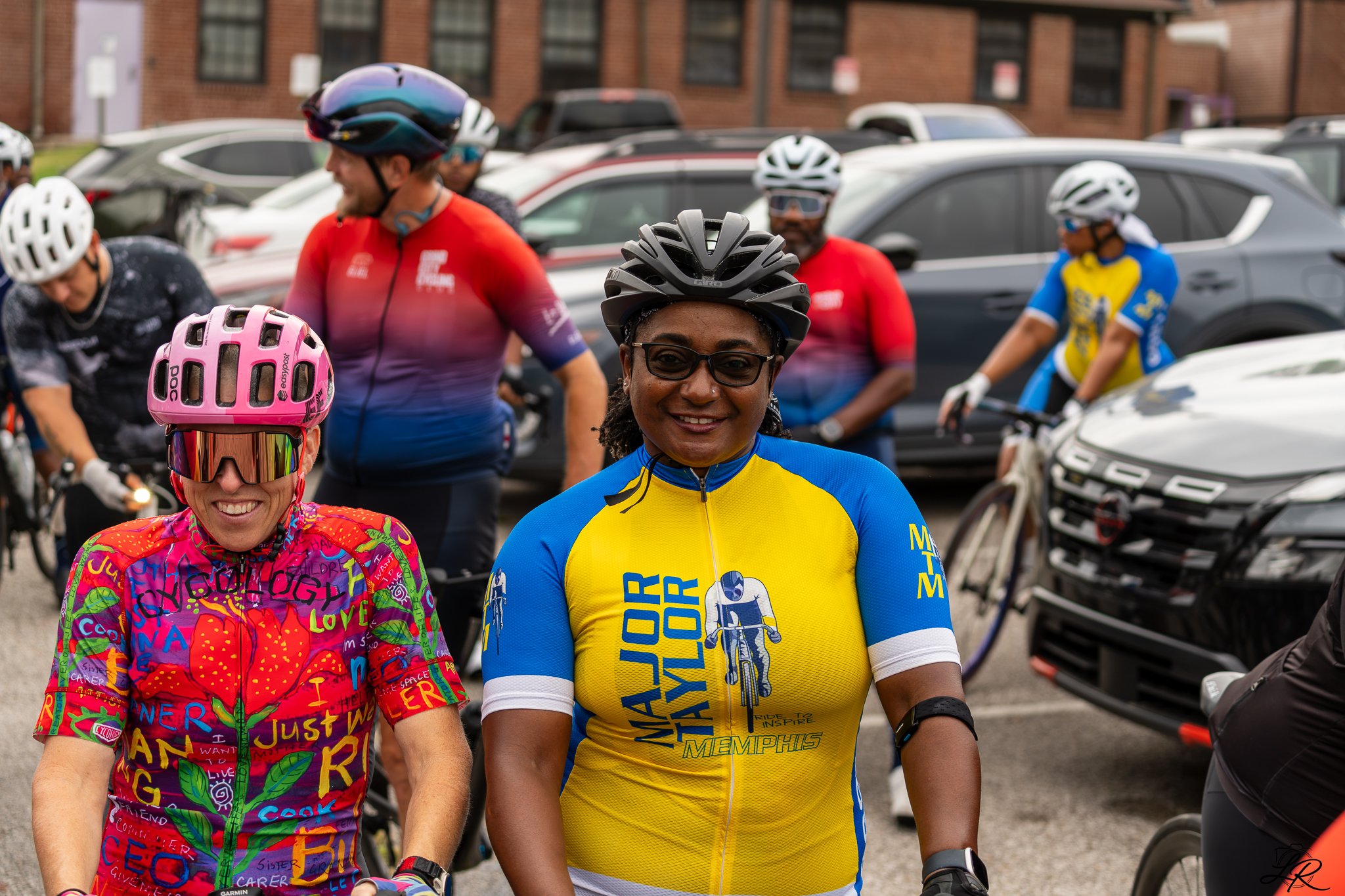 Group of smiling cyclists in colorful jerseys and helmets gathered outdoors, with parked cars and a brick building in the background.