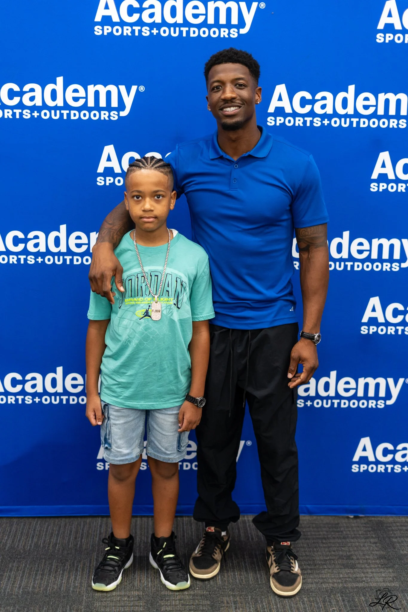 A young boy and an adult man standing together in front of a blue backdrop with 'Academy Sports + Outdoors' logo, both dressed in casual sportswear and smiling at the camera.