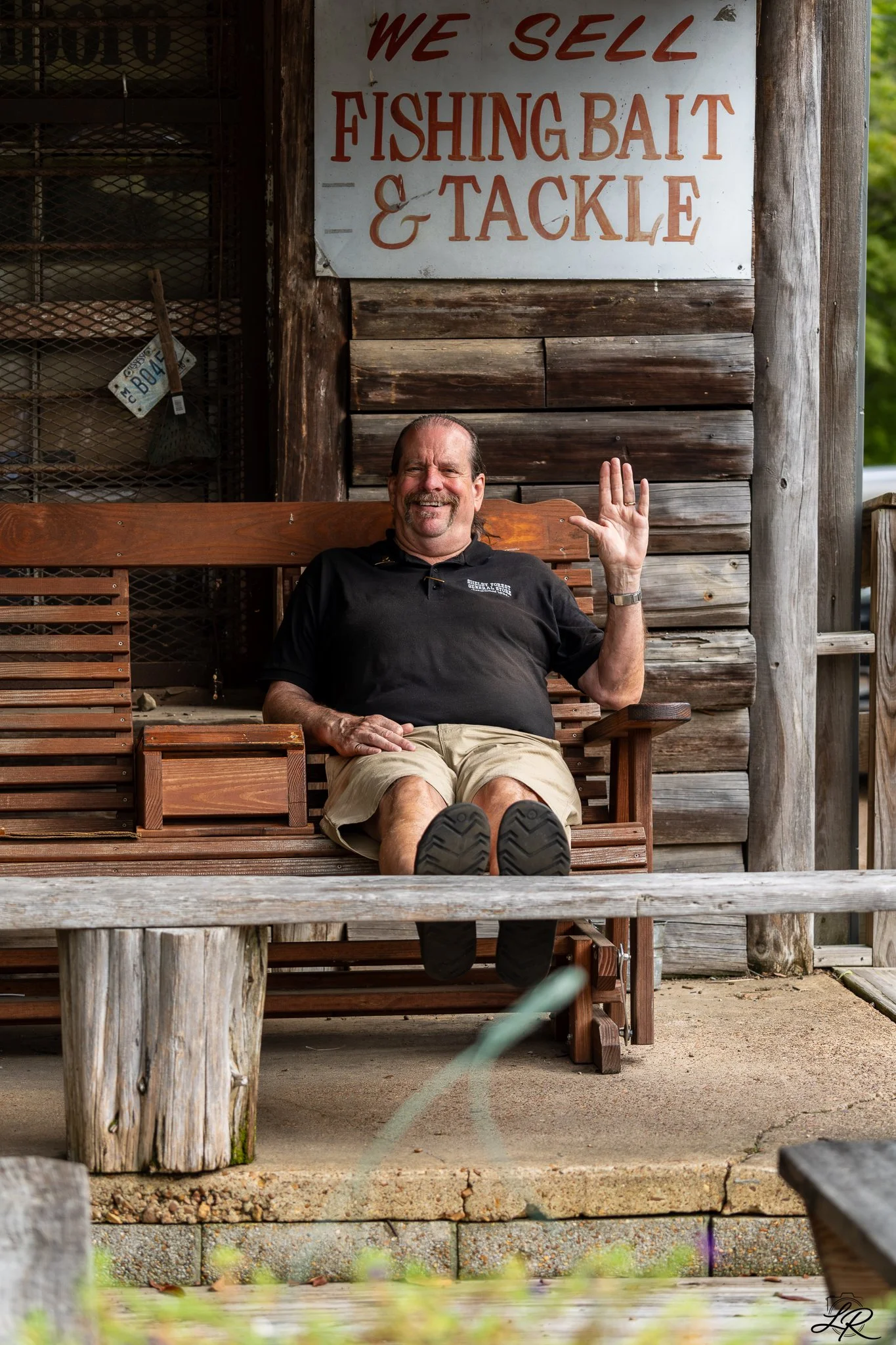 A man sits on a wooden bench on a porch, smiling and waving, with a white sign behind him that reads 'We Sell Fishing Bait & Tackle'.
