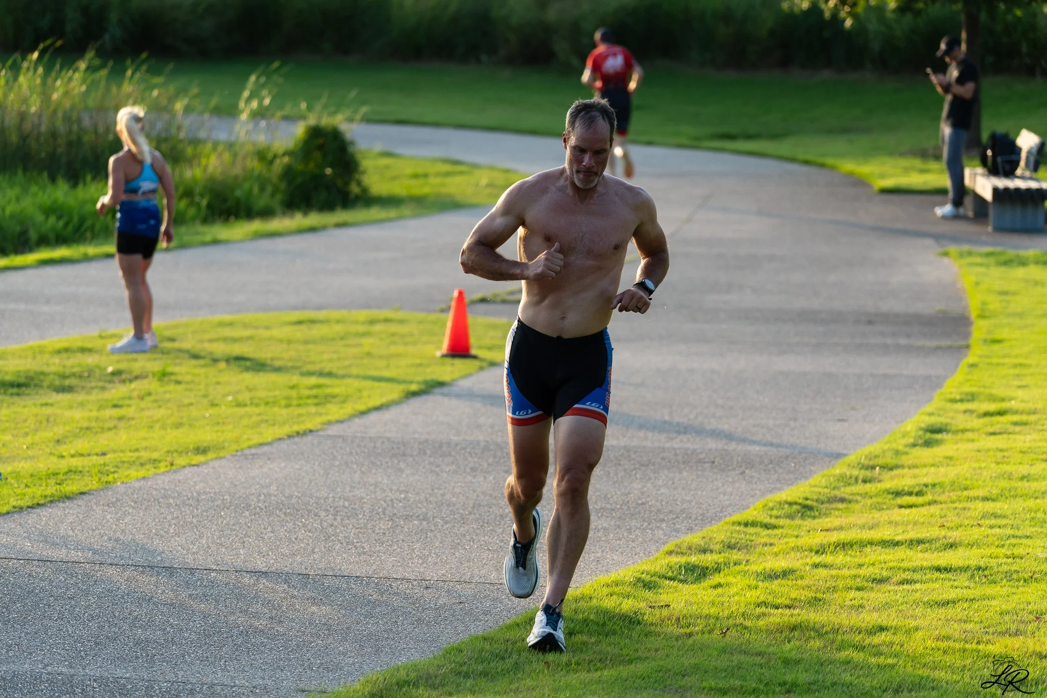 A shirtless man running along a paved path in a park, with a cone marker nearby, accompanied by a woman in athletic wear and other people in the background, in green surroundings.