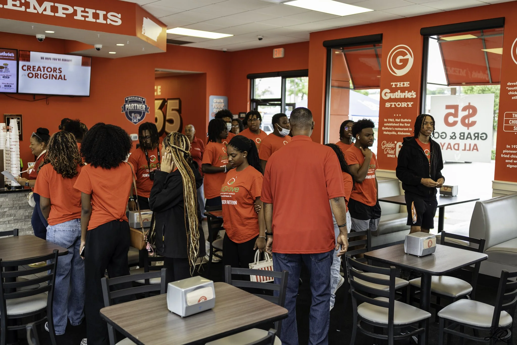 Inside a fast-food restaurant with red walls and several people standing in line, some wearing red shirts. There are multiple televisions on the wall, one displaying the words "CREATORS ORIGINAL" and another displaying "THE GUTHRIE'S STORY." There ar