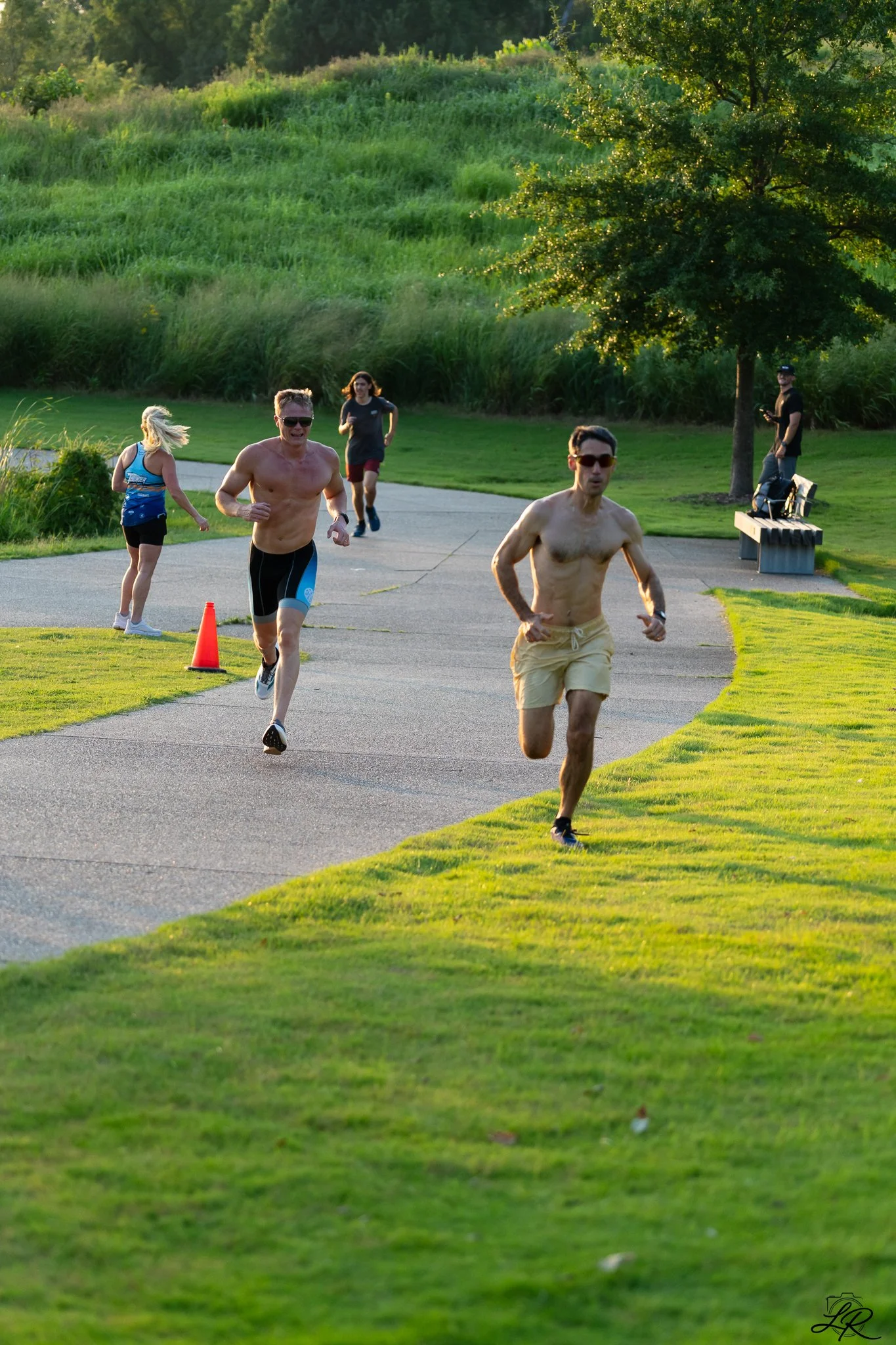 People running on a paved path in a park during sunset, with trees and lush green grass in the background.