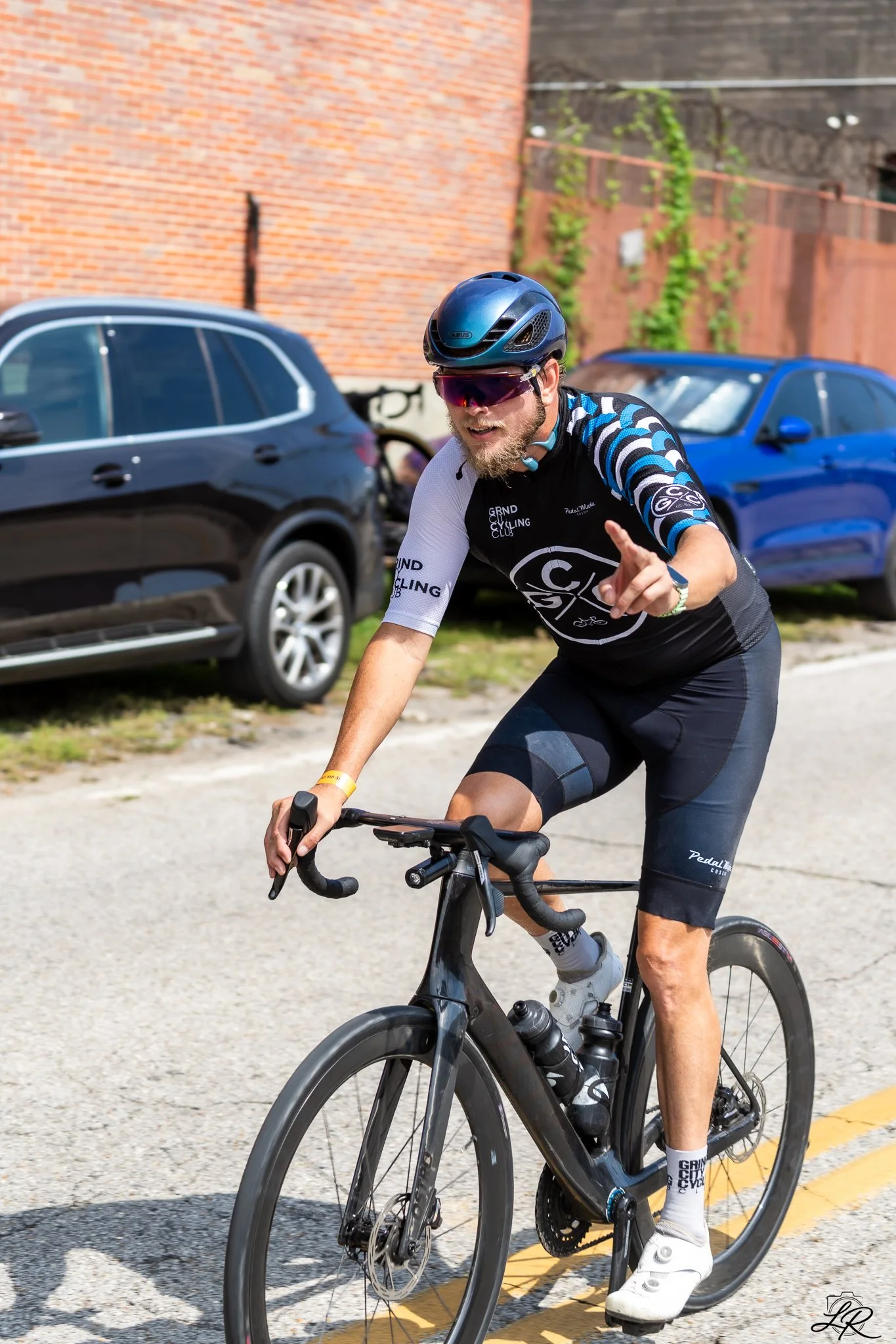 A man riding a black road bicycle on a street, wearing a blue helmet, sunglasses, cycling jersey, shorts, and white cycling shoes, with cars and a brick wall in the background.