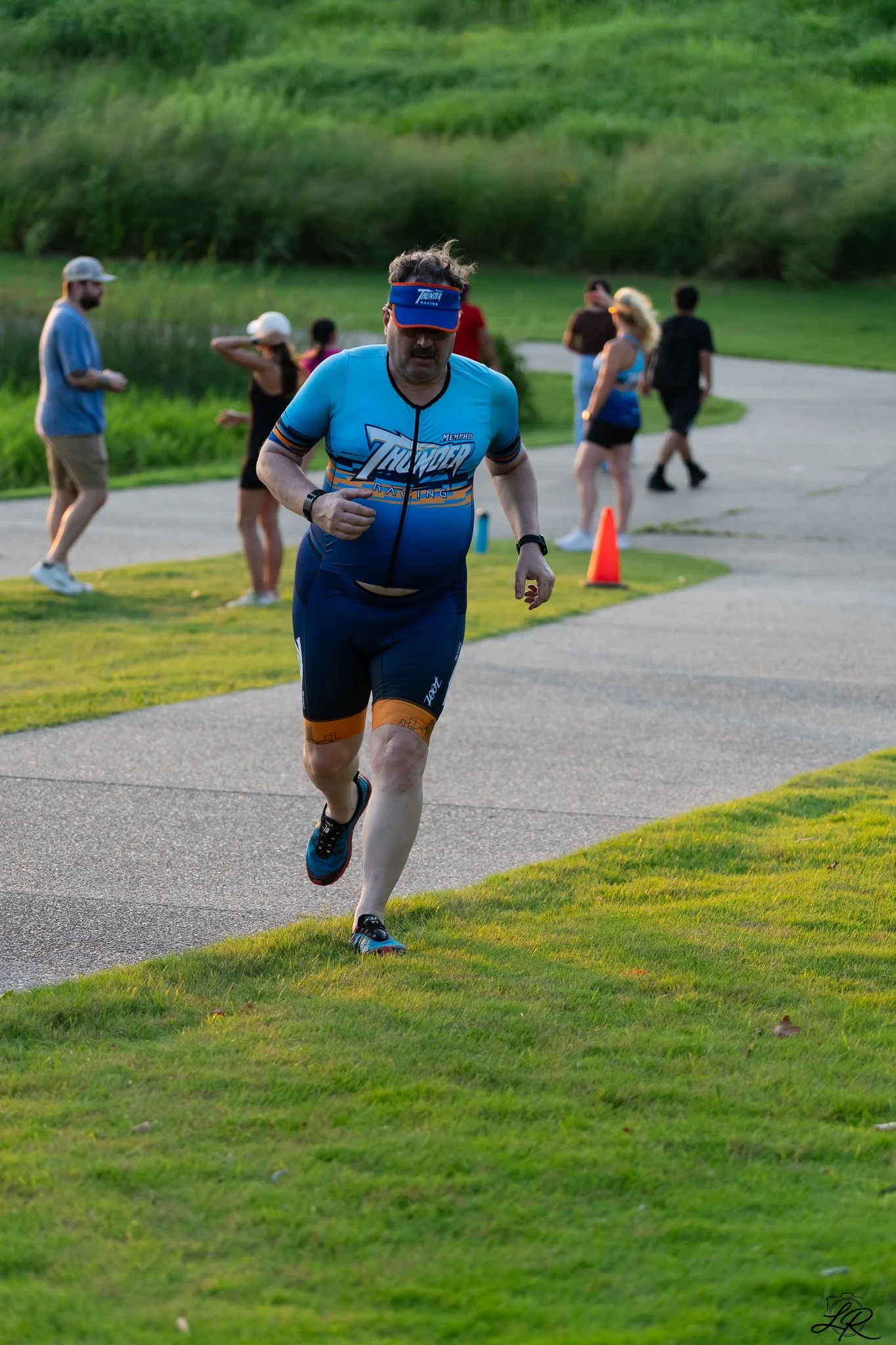 A man running on a paved path in a park with other people standing and walking around in the background, surrounded by greenery.
