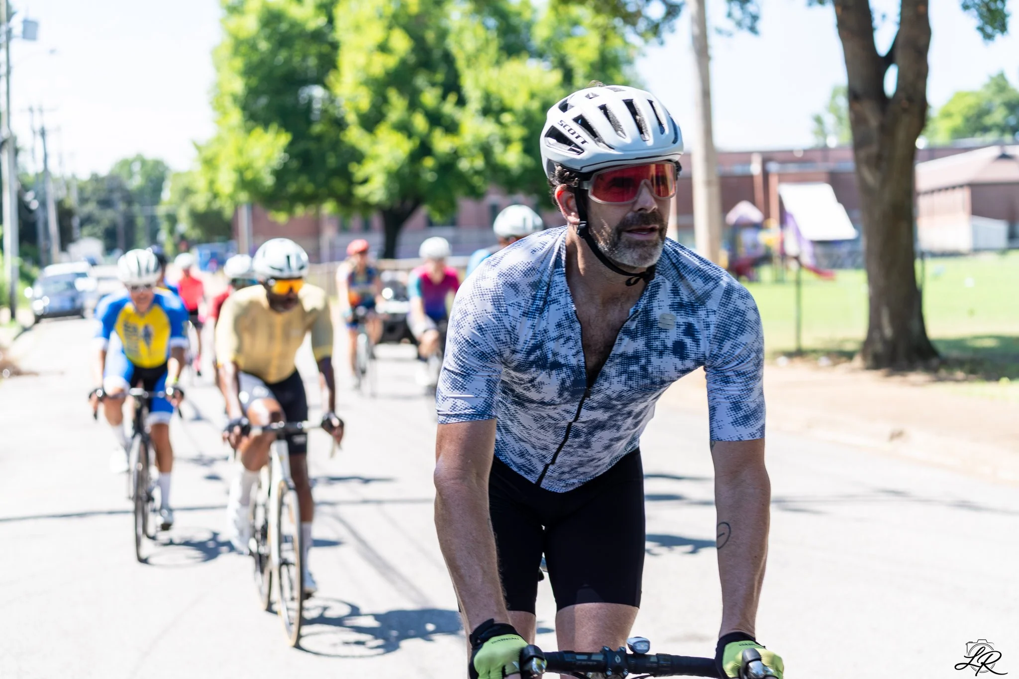 A man riding a bicycle in a cycling event, wearing a white helmet, red sunglasses, and a patterned cycling jersey, with other cyclists and trees in the background on a sunny day.