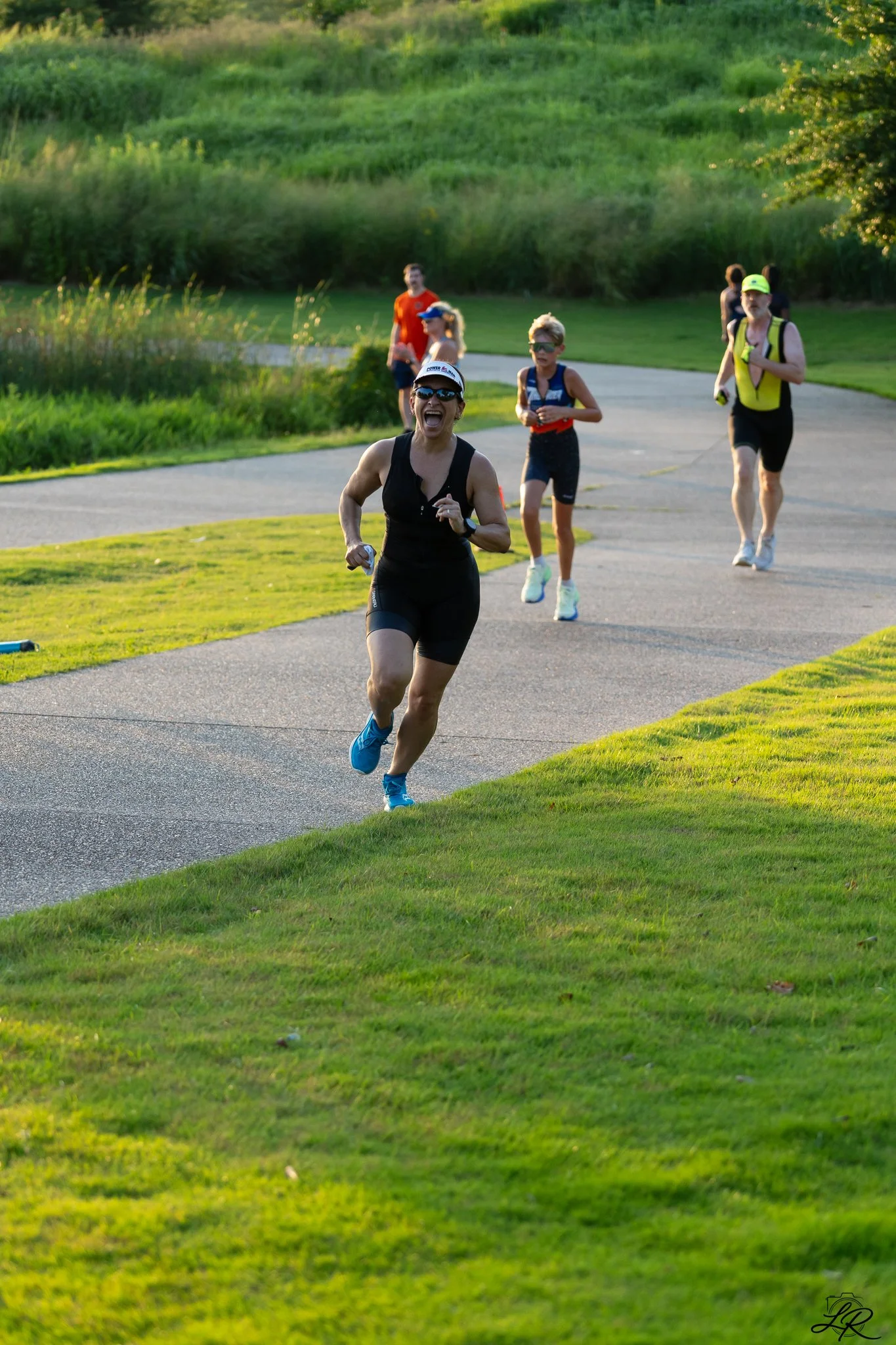 A group of four runners participating in an outdoor race on a paved path surrounded by lush green grass and trees. The woman in the front is wearing a black outfit, sunglasses, and a cap, smiling and looking energetic. The other runners are a mix of 
