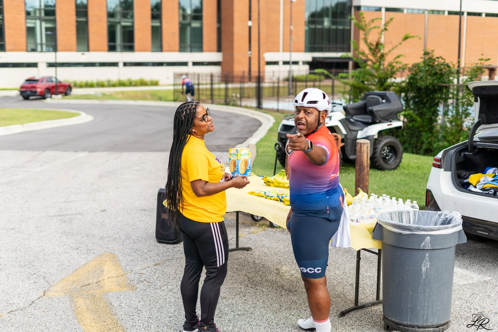 A woman in a yellow shirt and glasses is talking to a man dressed in cycling gear, including a helmet, next to a table with water bottles and snacks at an outdoor event, near a parking lot and a modern brick building.