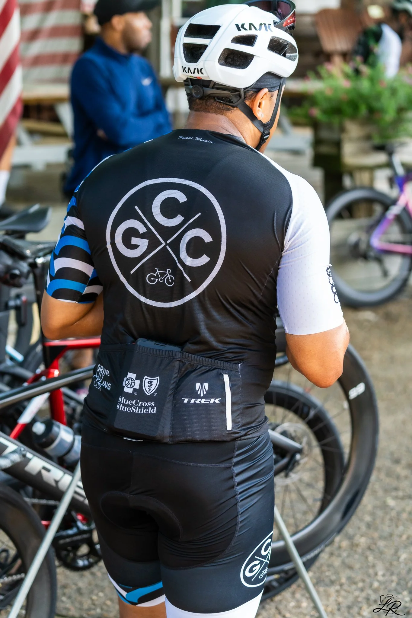 A cyclist wearing a white helmet and a black cycling jersey with various sponsor logos, standing among other bicycles and people.