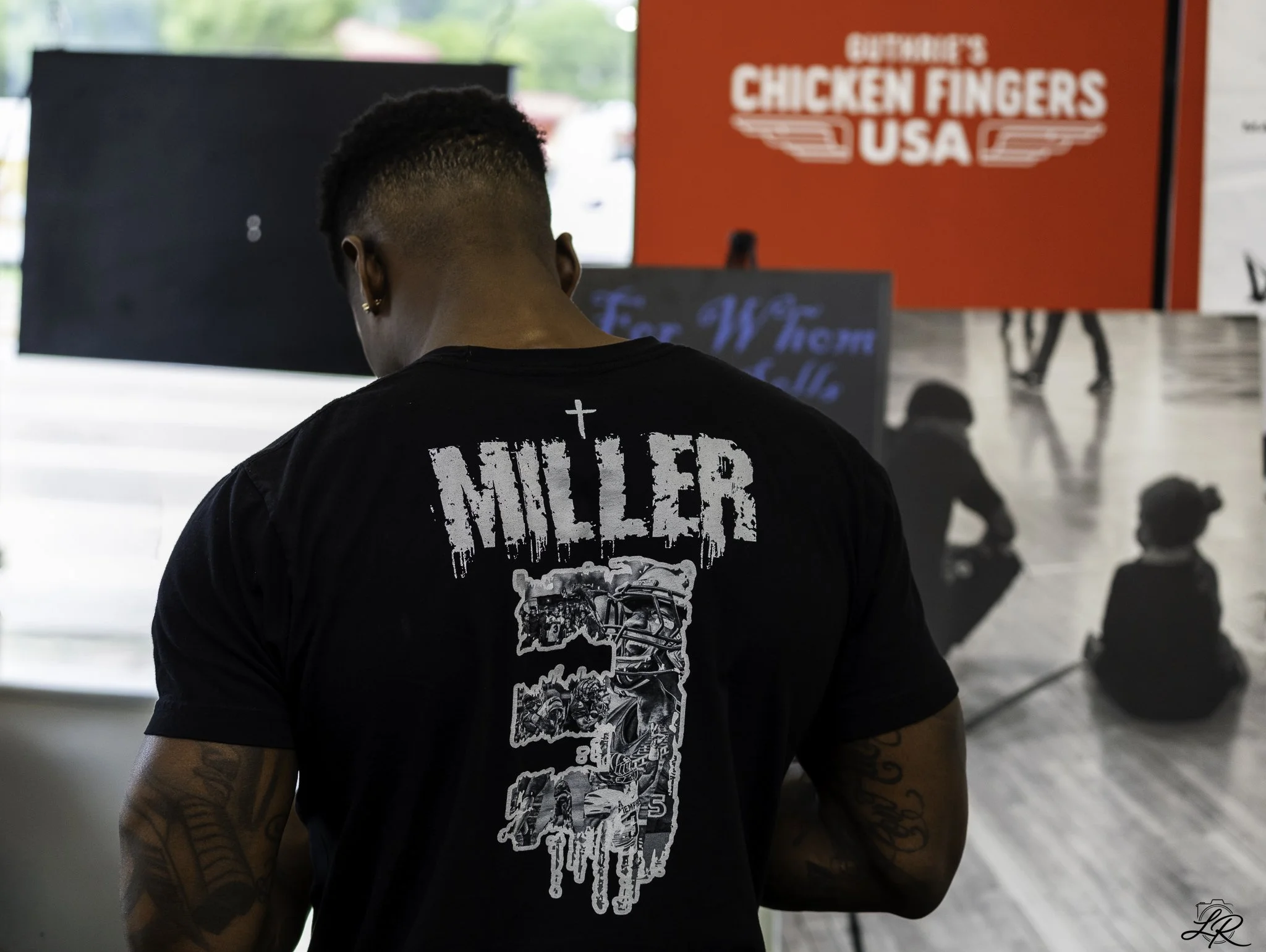 Person with tattoos on arms wearing a black shirt with a graphic and the word 'KILLER' on the back, standing in front of red and black signs, including a sign that says 'CHICKEN FINGERS USA'.