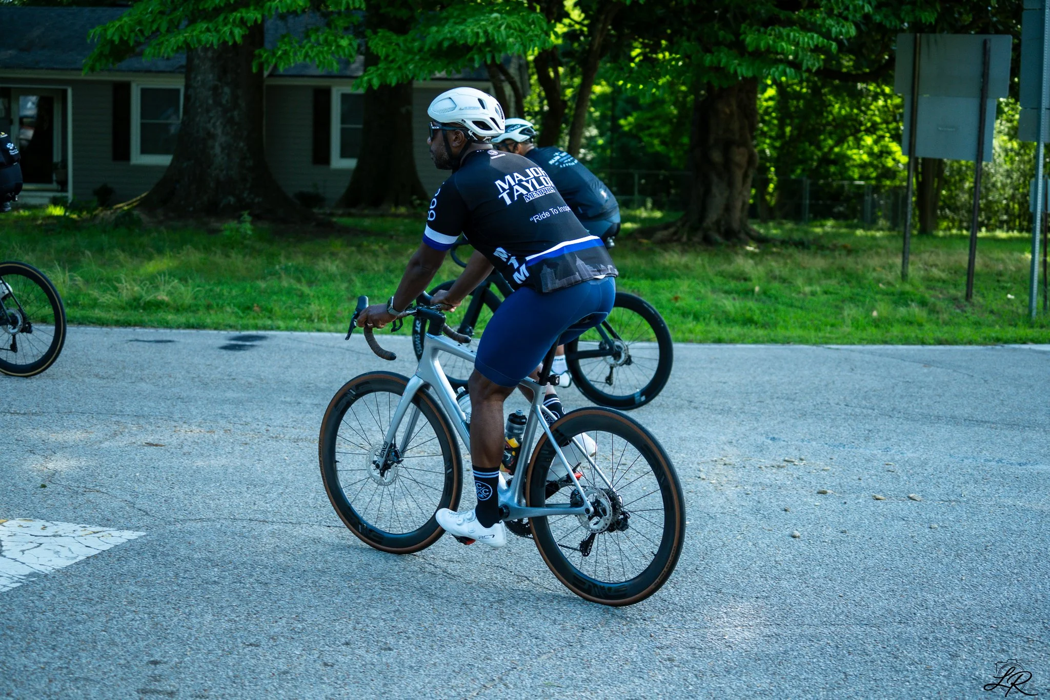 Cyclist wearing a black jersey and helmet riding a bicycle on a paved road with trees and houses in the background.