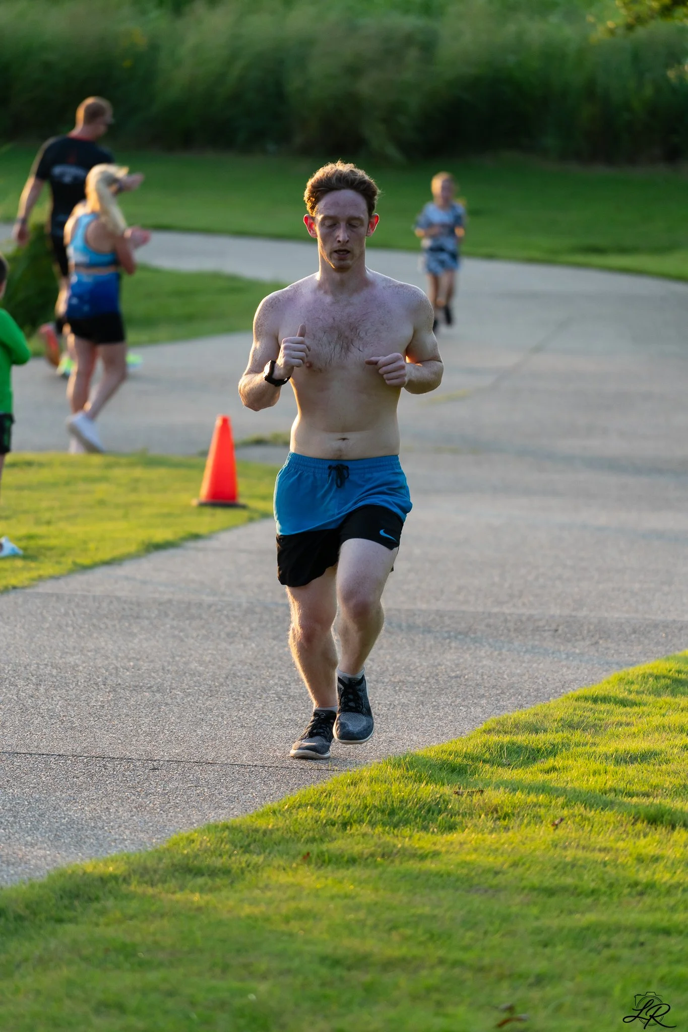 A man running on a paved path outdoors during sunset, wearing blue shorts, black running shoes, and a fitness tracker on his left wrist, with a grassy area and trees in the background.