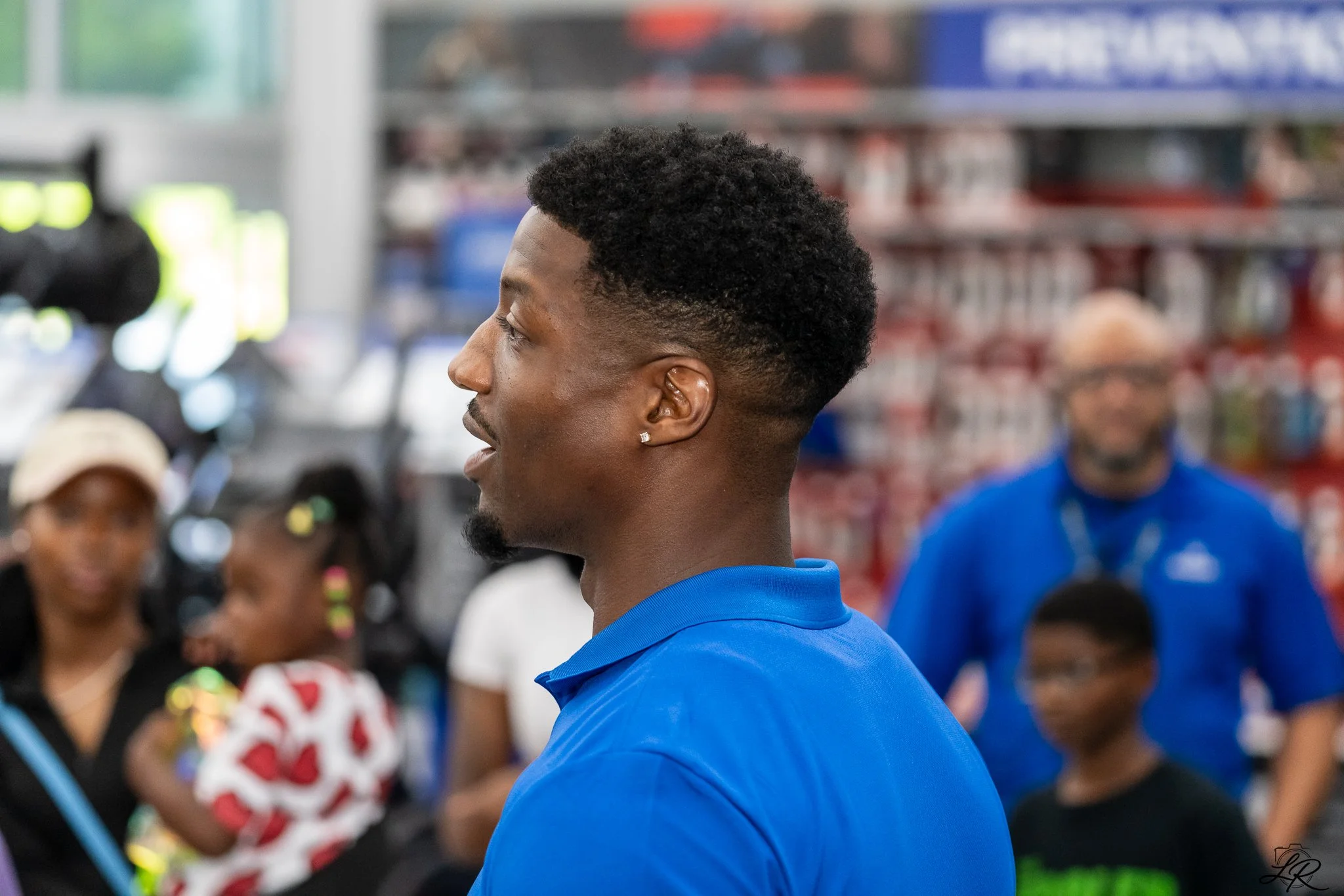 A young man with a short curly hairstyle, earrings, wearing a blue sports jersey, standing in profile, in what appears to be a store with blurred shelves and people in the background.