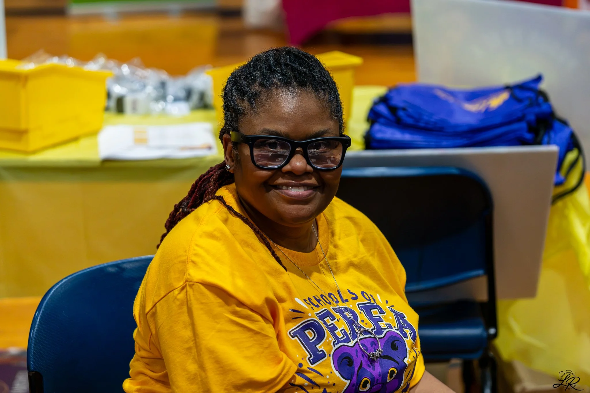 A smiling woman with glasses sitting in a room with yellow tables and boxes, wearing a yellow T-shirt.