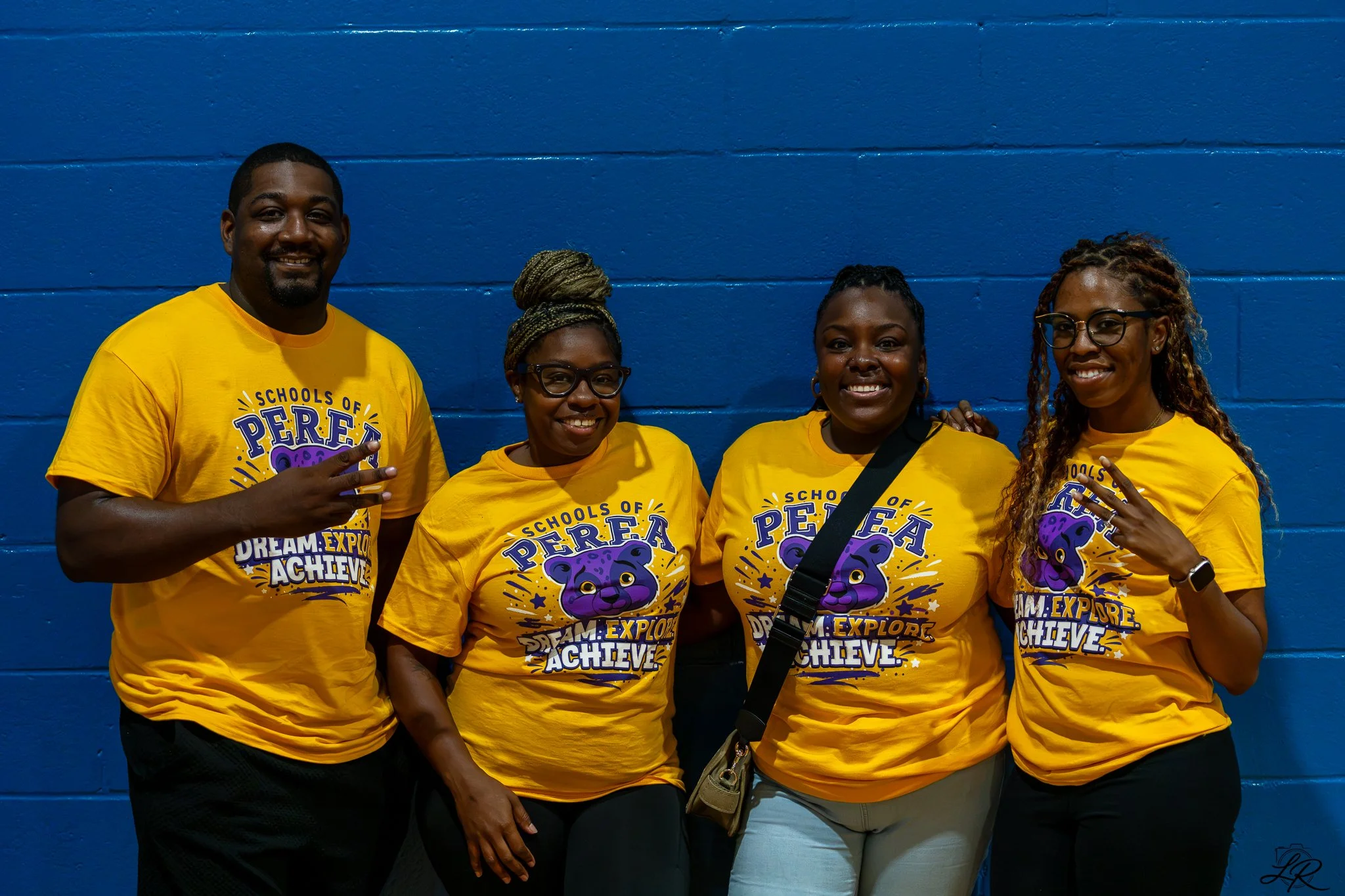 Four individuals standing against a blue wall, all wearing matching yellow T-shirts with a bear graphic and the words 'Schools of PERA, Explore, Dream, Achieve.' They are smiling and posing for the photo.