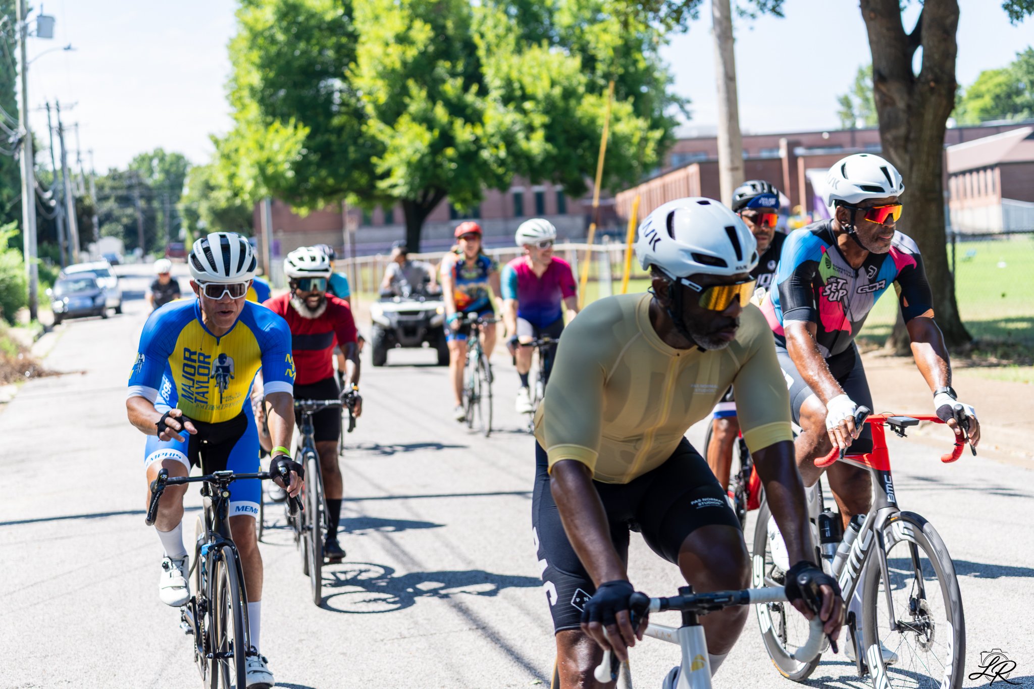 A group of people riding bicycles together on a sunny day in a suburban neighborhood, wearing helmets and sports gear.