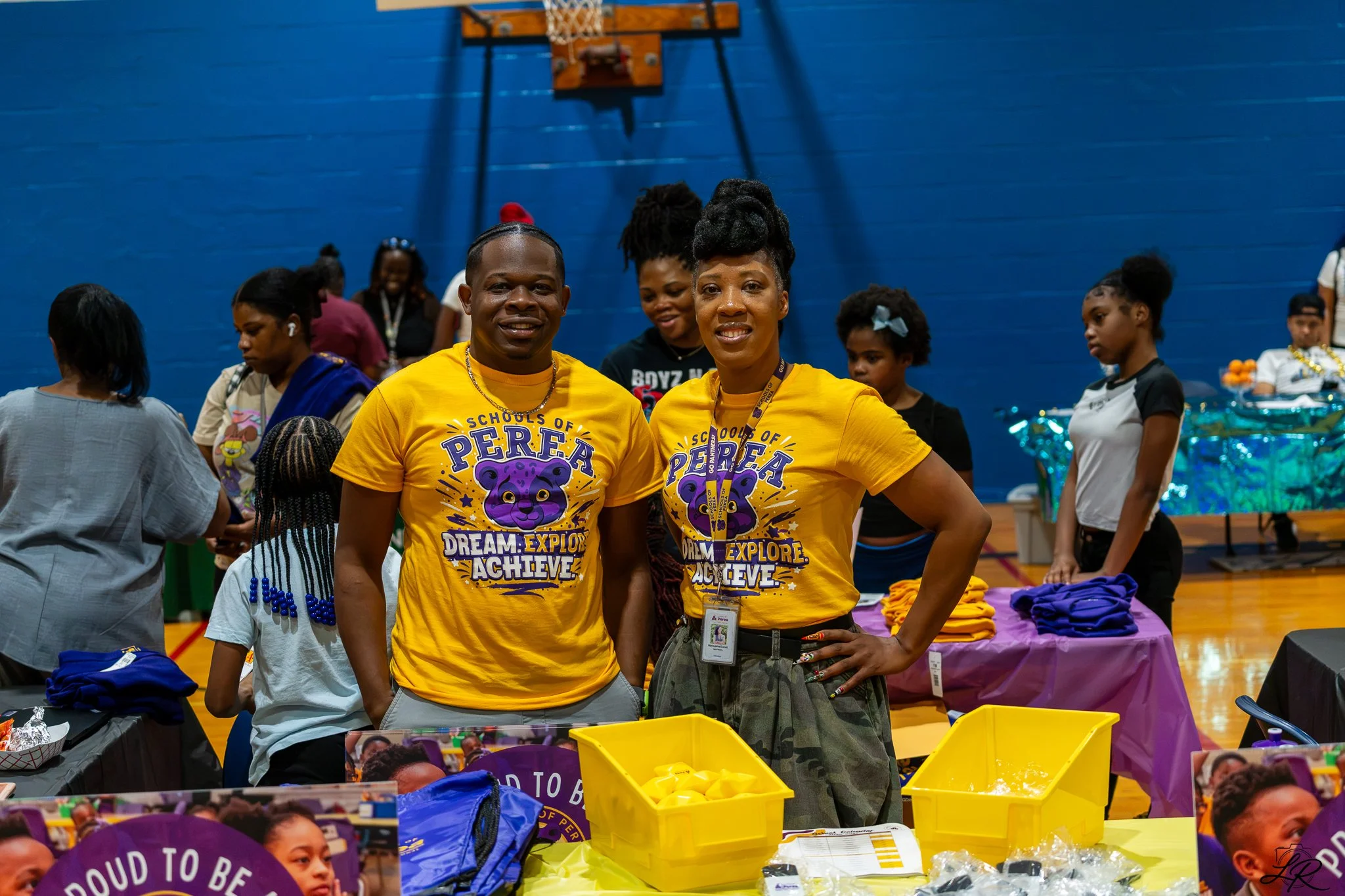 Two women wearing yellow shirts with a purple bear logo, standing in a gymnasium, surrounded by children and other people, at an event promoting education and achievement, with tables and yellow bins in the foreground.