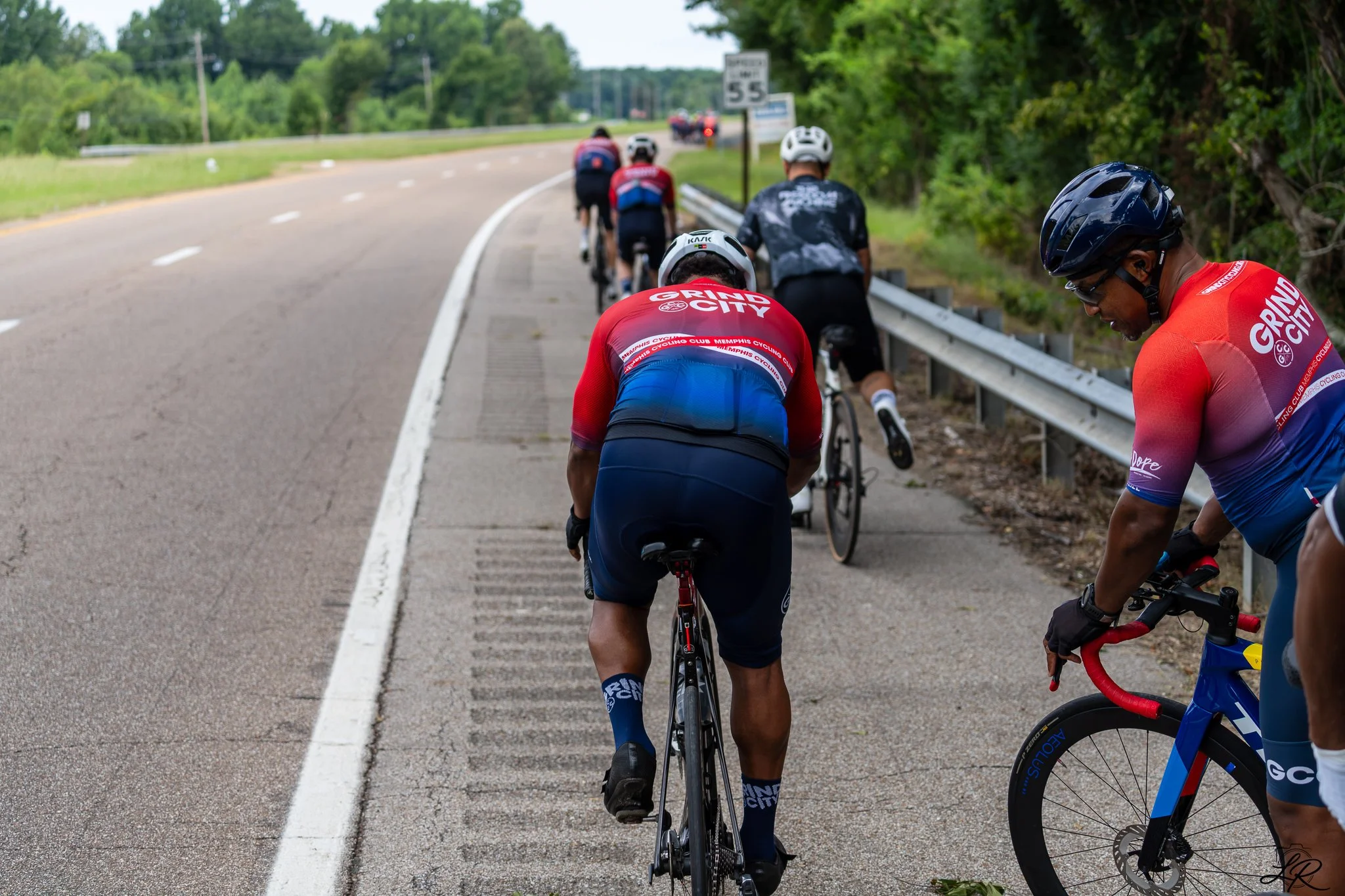 Cyclists in athletic gear riding bicycles along a rural road with trees and a guardrail, some standing and some riding.