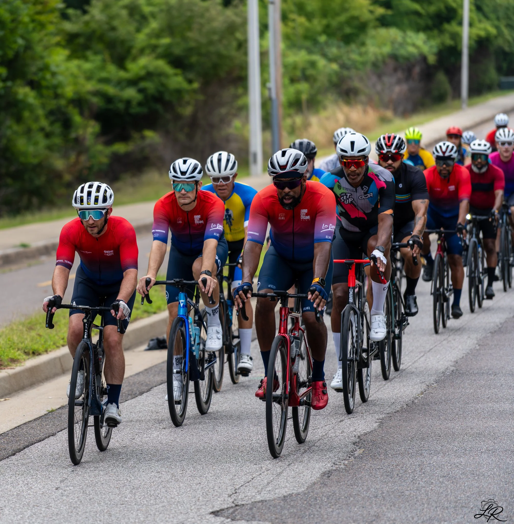 A group of cyclists riding on a paved road surrounded by lush green trees, all wearing helmets and colorful cycling gear.