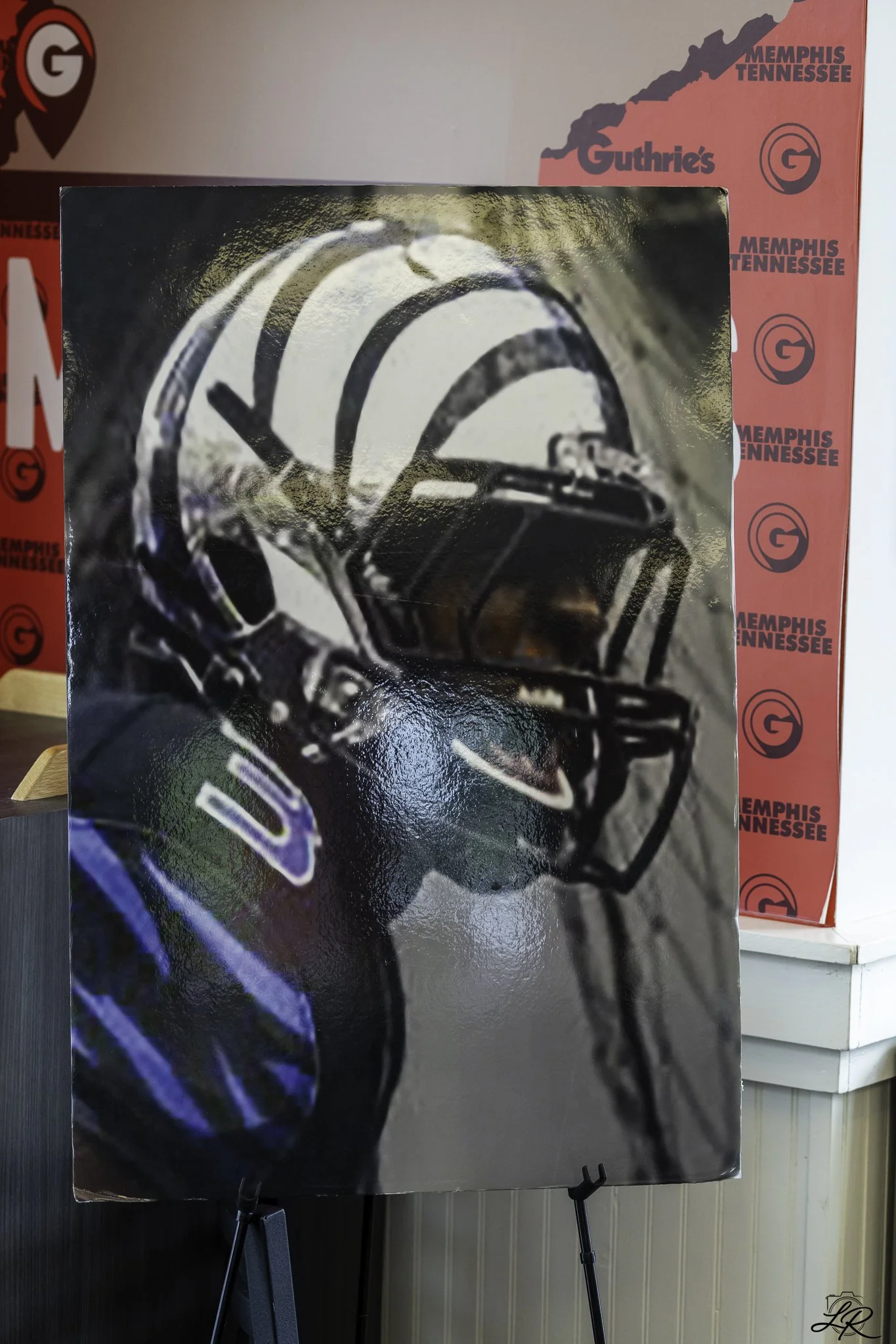 Poster featuring a close-up image of a football helmet with a black face mask, predominantly black and white with hints of blue and purple, displayed on an easel in front of a red and white wall with Memphis Tennessee branding.