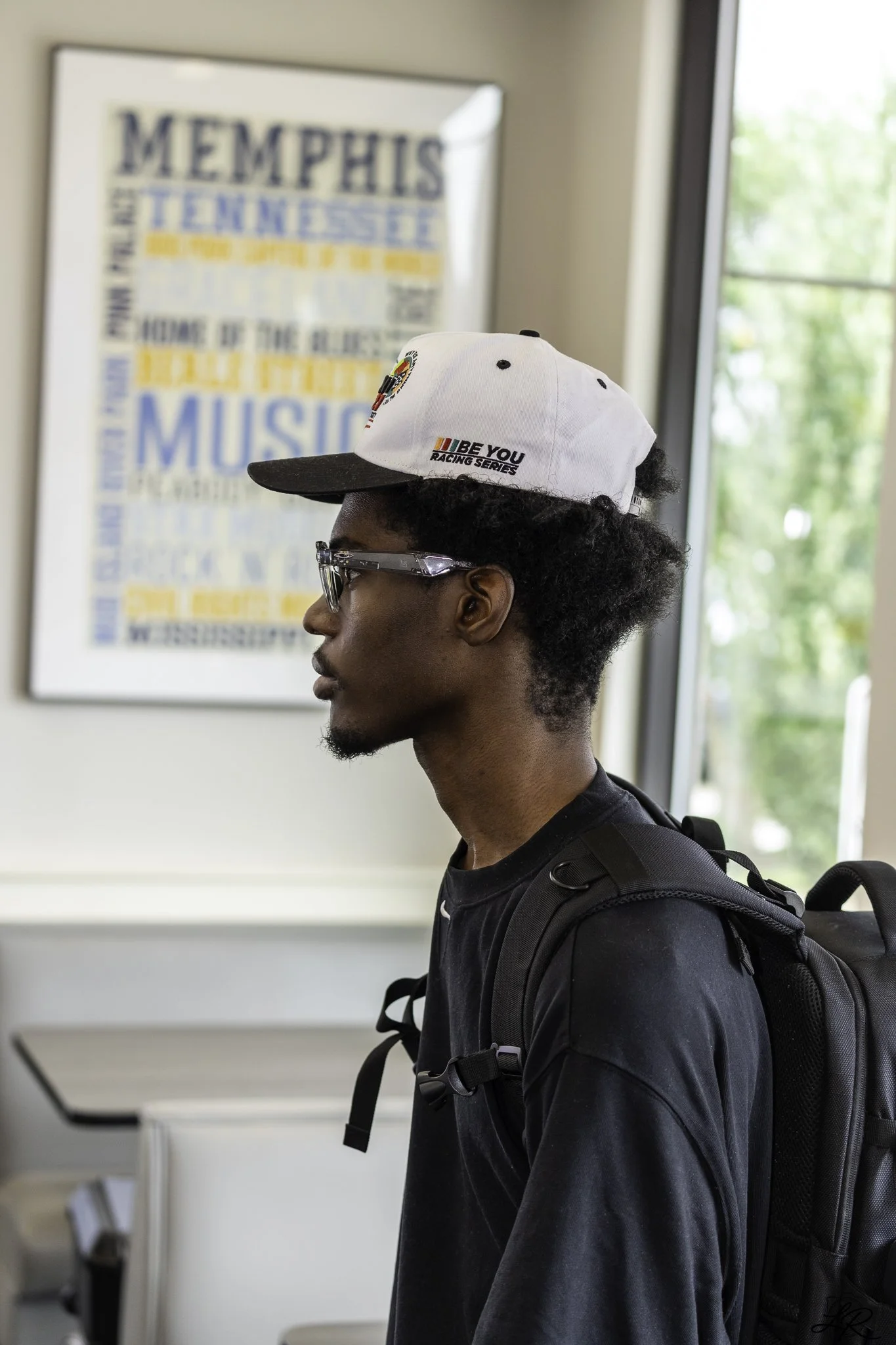 Profile of a young man with glasses, wearing a white cap with logos, a black shirt, and a backpack, standing indoors near a window with greenery outside and a blurry poster in the background.