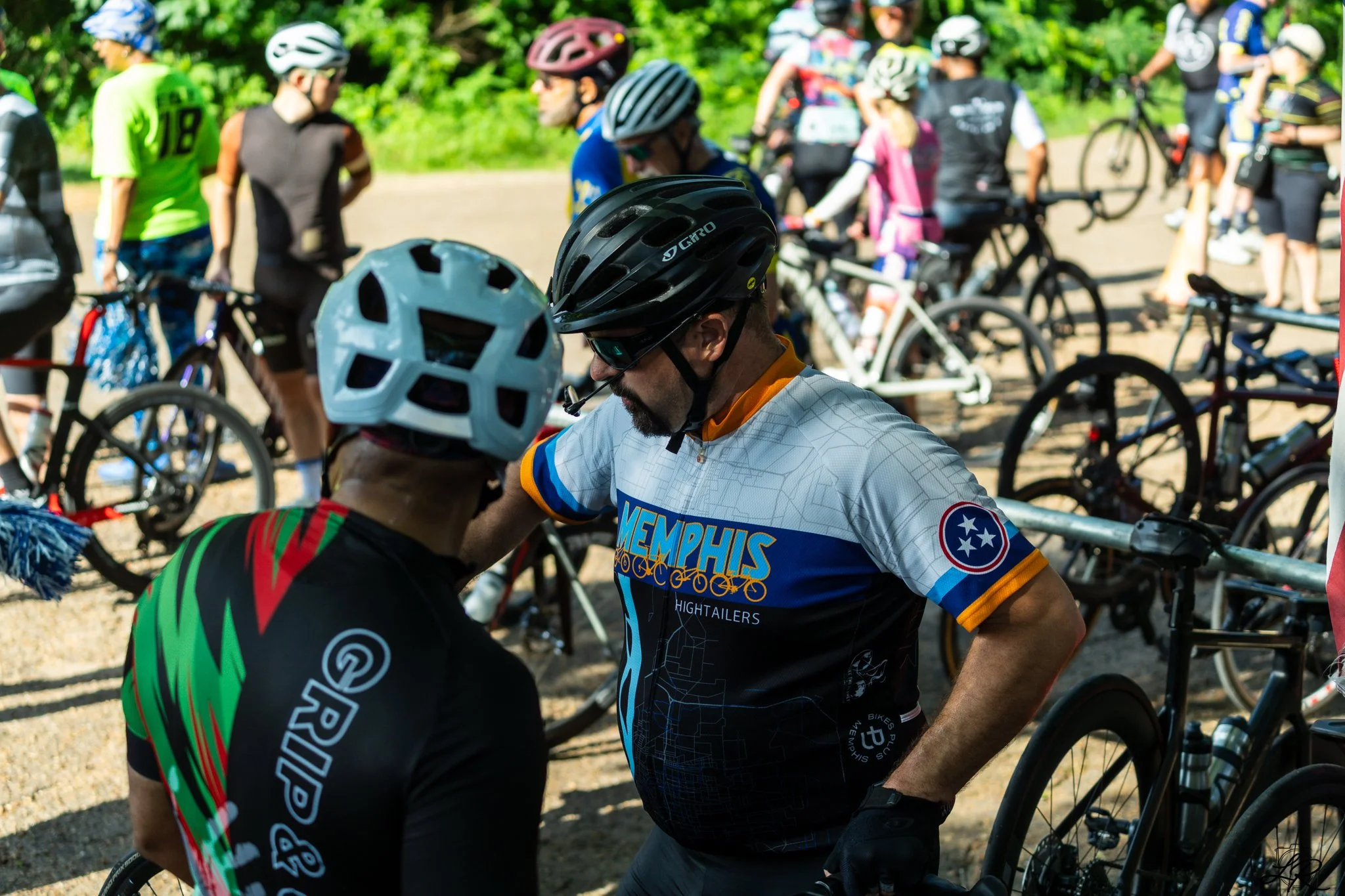 Cyclists gathering outdoors, some wearing helmets and sports attire, with bicycles parked nearby and greenery in the background.