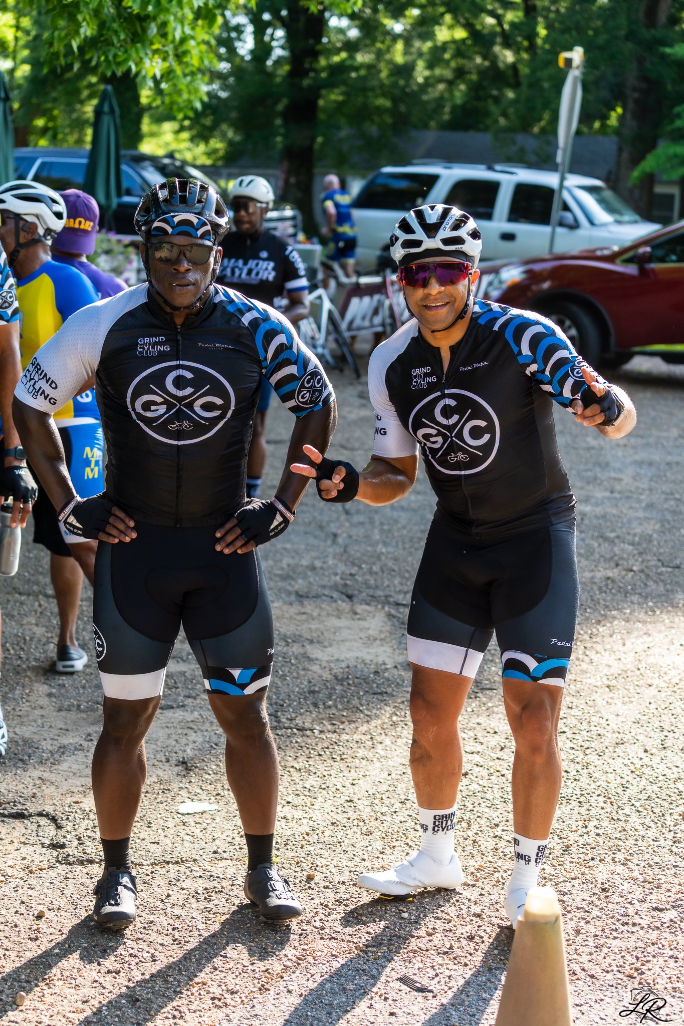 Two male cyclists in matching black, white, and blue cycling jerseys and shorts, wearing helmets and sunglasses, standing outdoors on a gravel path with parked cars and trees in the background.