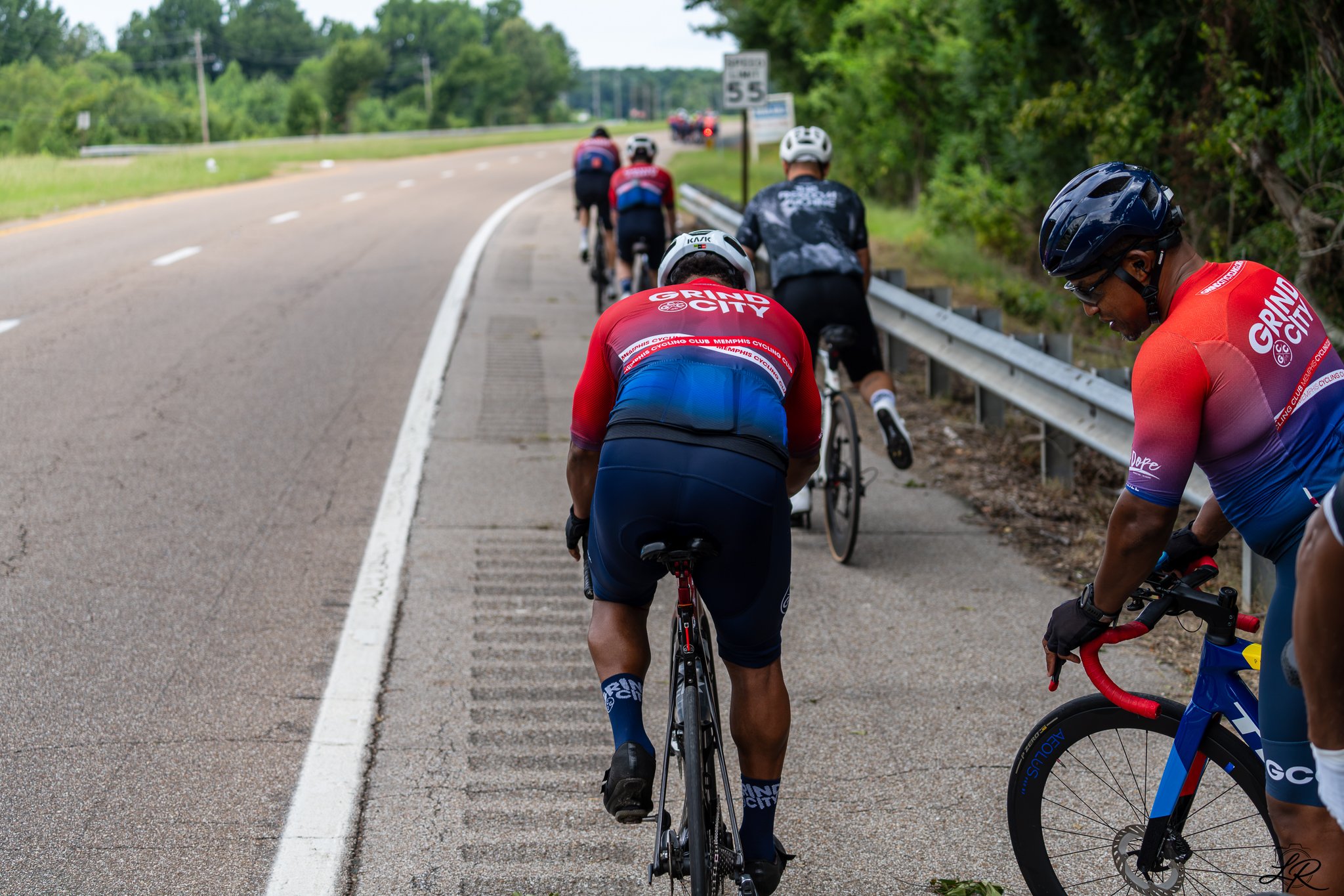 Group of cyclists wearing helmets and cycling jerseys, riding on a road with a guardrail on the right side, surrounded by green trees and a grassy area, with a speed limit sign visible in the background.