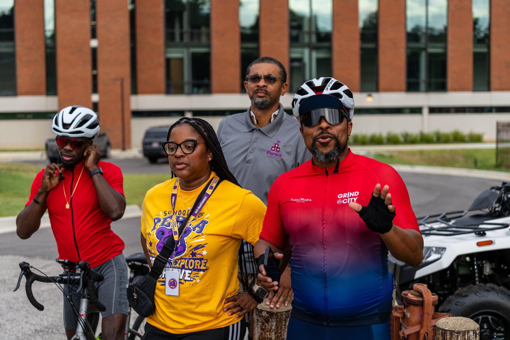 Group of four diverse people outdoors, some wearing bike helmets, standing with bicycles, in front of a modern brick building.